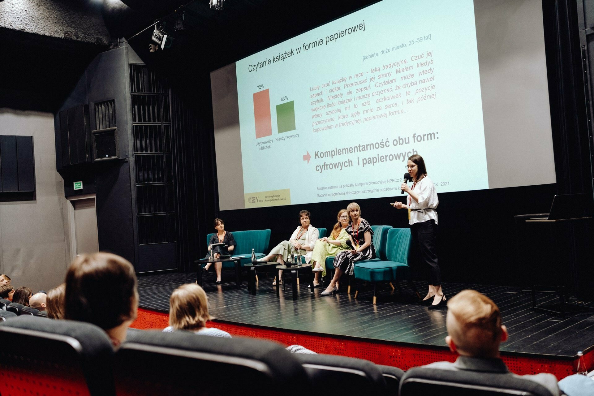 A group of five people sit on a stage in front of a screen displaying a presentation. One person stands and speaks into a microphone, while the others sit. The seated audience watches the presentation given in Polish in what resembles a lecture hall. A photographer from Warsaw could provide an excellent photo essay of the event.   
