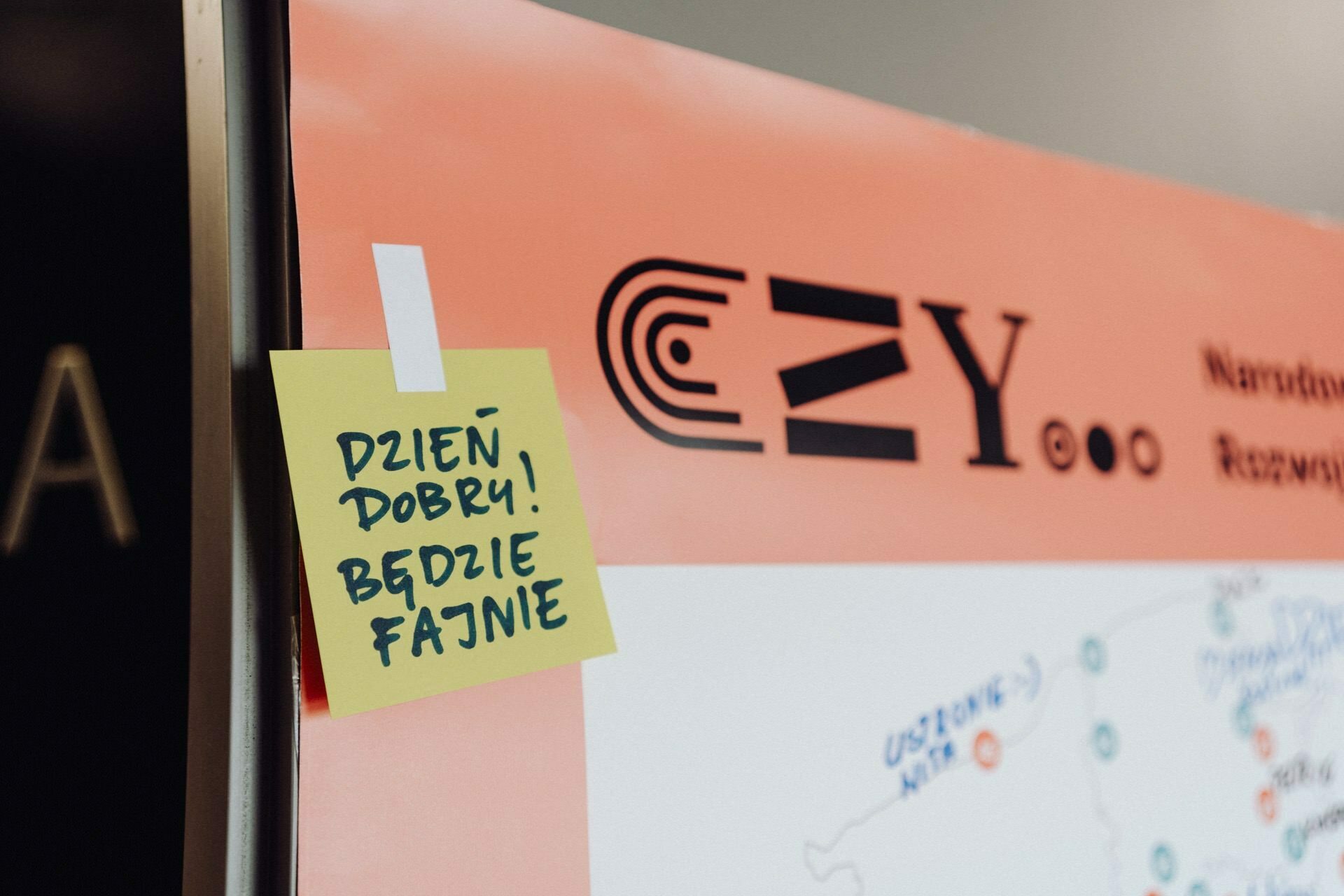 Close-up of the board with a sticky note in Polish "DAWN GOOD! WILL BE FUN!", in front of the section with the text "EZ. Y" and other partially visible elements. The combination of text and graphics suggests an informational or presentation display, ideal for photographing events where details can be captured.   