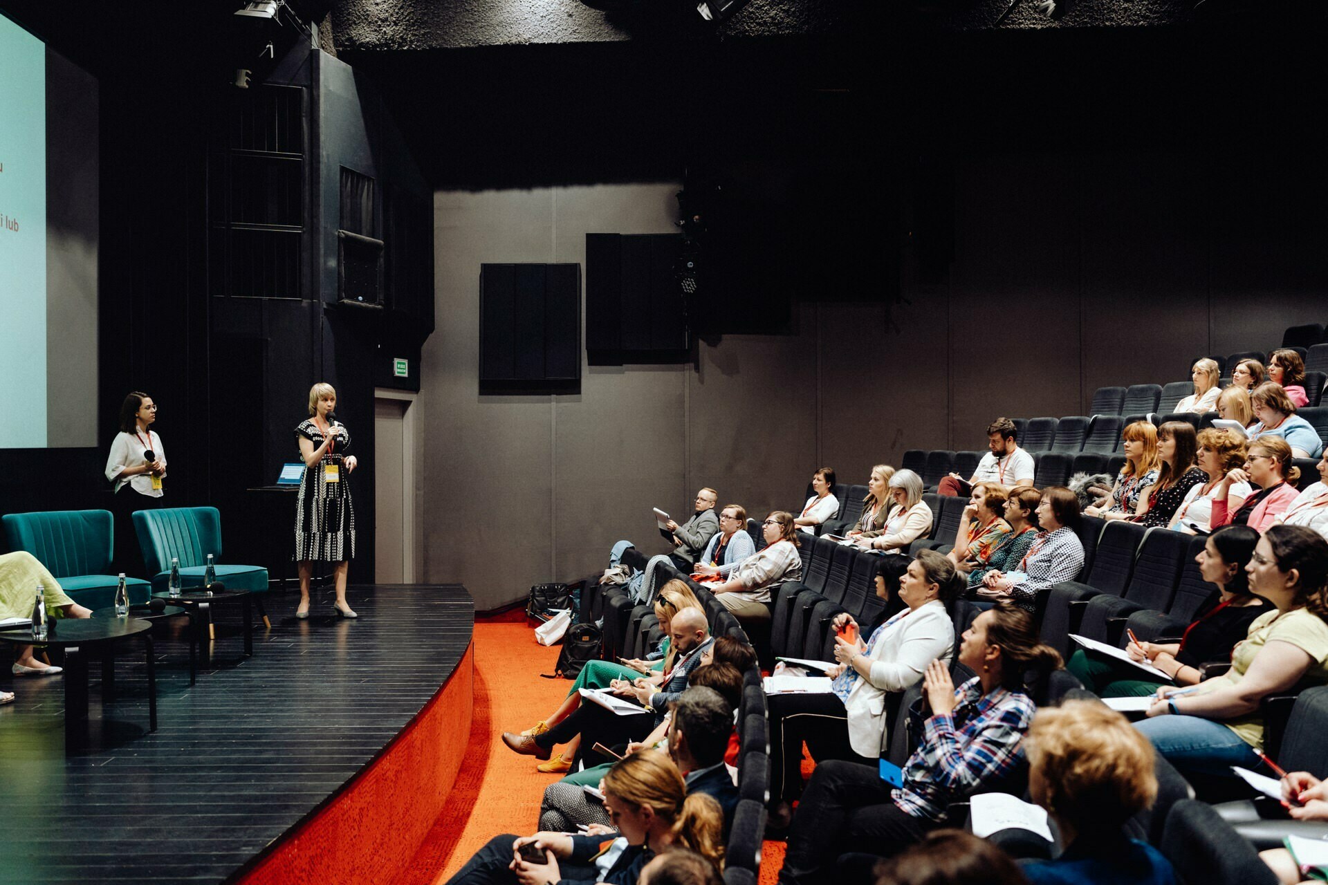 The speaker stands on a stage in front of an audience in a dimly lit auditorium. The audience sits at desks with notepads and laptops and listens intently. A photographer at the event moves near the speaker's podium, capturing the moments. The room has a modern decor with dark walls and green chairs.   