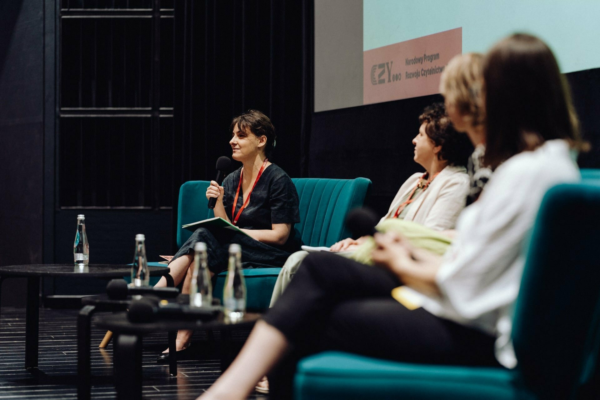 Four people sit in a staged panel discussion. One woman speaks into a microphone, two others listen intently. In the foreground is another person, out of focus. They all have water bottles and are wearing conference lanyards. This scene captures the essence of an engaging event taking place in Warsaw, perfect for an event photographer warsaw.    