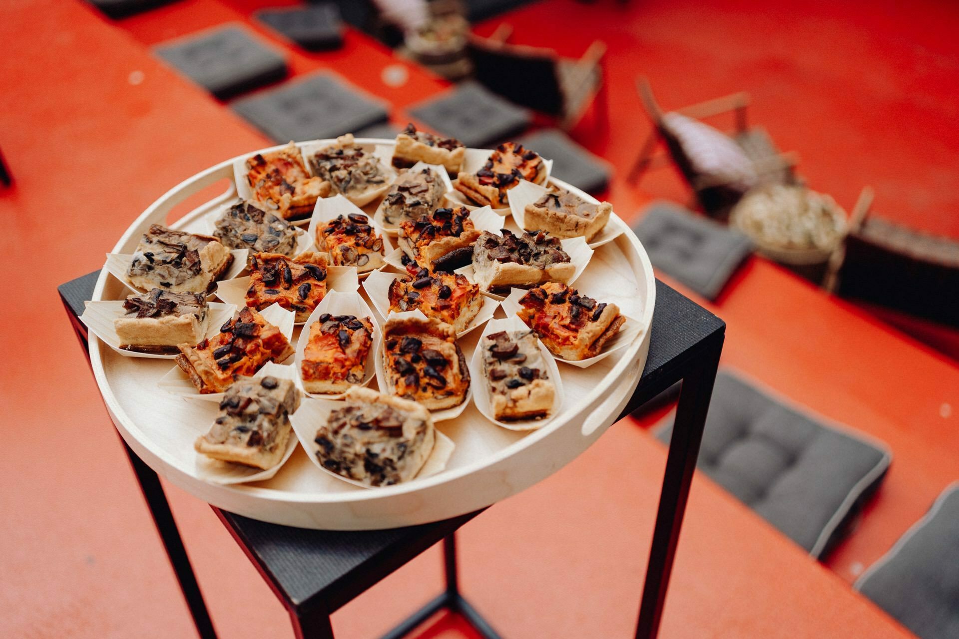 A round tray filled with a variety of small appetizers stands on a black stand. Each appetizer is served in a small, individual boat-shaped dish. The backdrop, reminiscent of a photo conference, has a red floor and scattered seating areas, including pillows and chairs.  