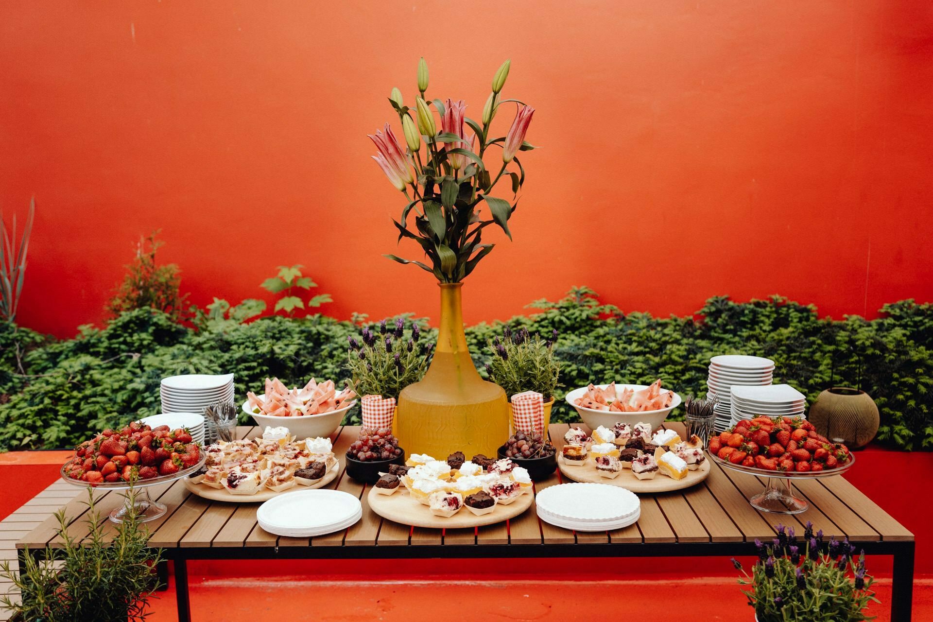 On a wooden table set outside against an orange wall are various desserts, bowls of strawberries and stacks of plates. The centerpiece is a tall yellow vase with pink flowers. Captured by a photographer from Warsaw, the scene shows lush greenery in the background.  