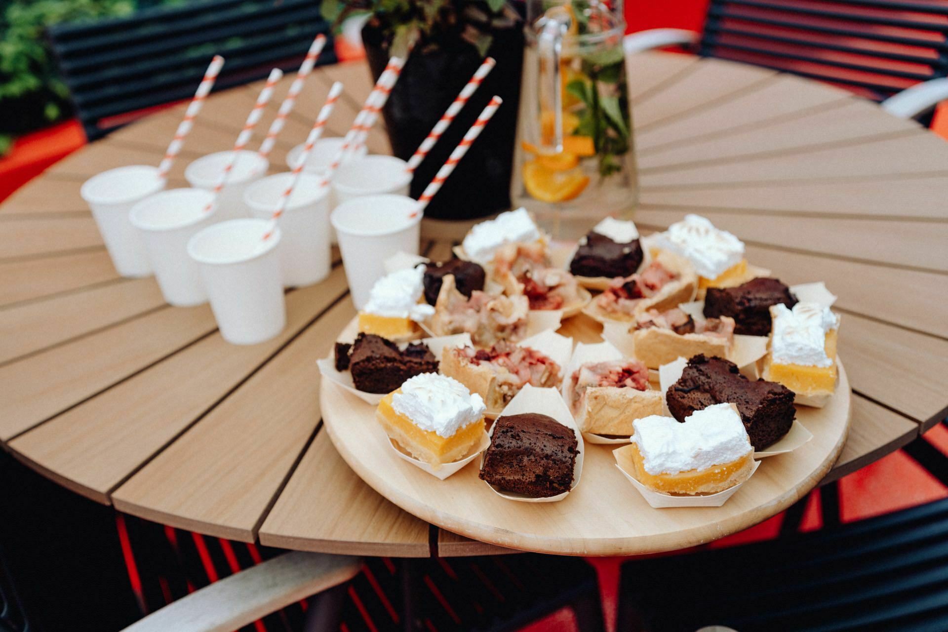 A plate of assorted desserts, including a brownie with whipped cream and other baked goods, is set on a round wooden table. Standing next to the plate are several white paper cups with red and white striped straws and a glass pitcher filled with citrus-infused water - a photogenic scene for any Warsaw photographer. 