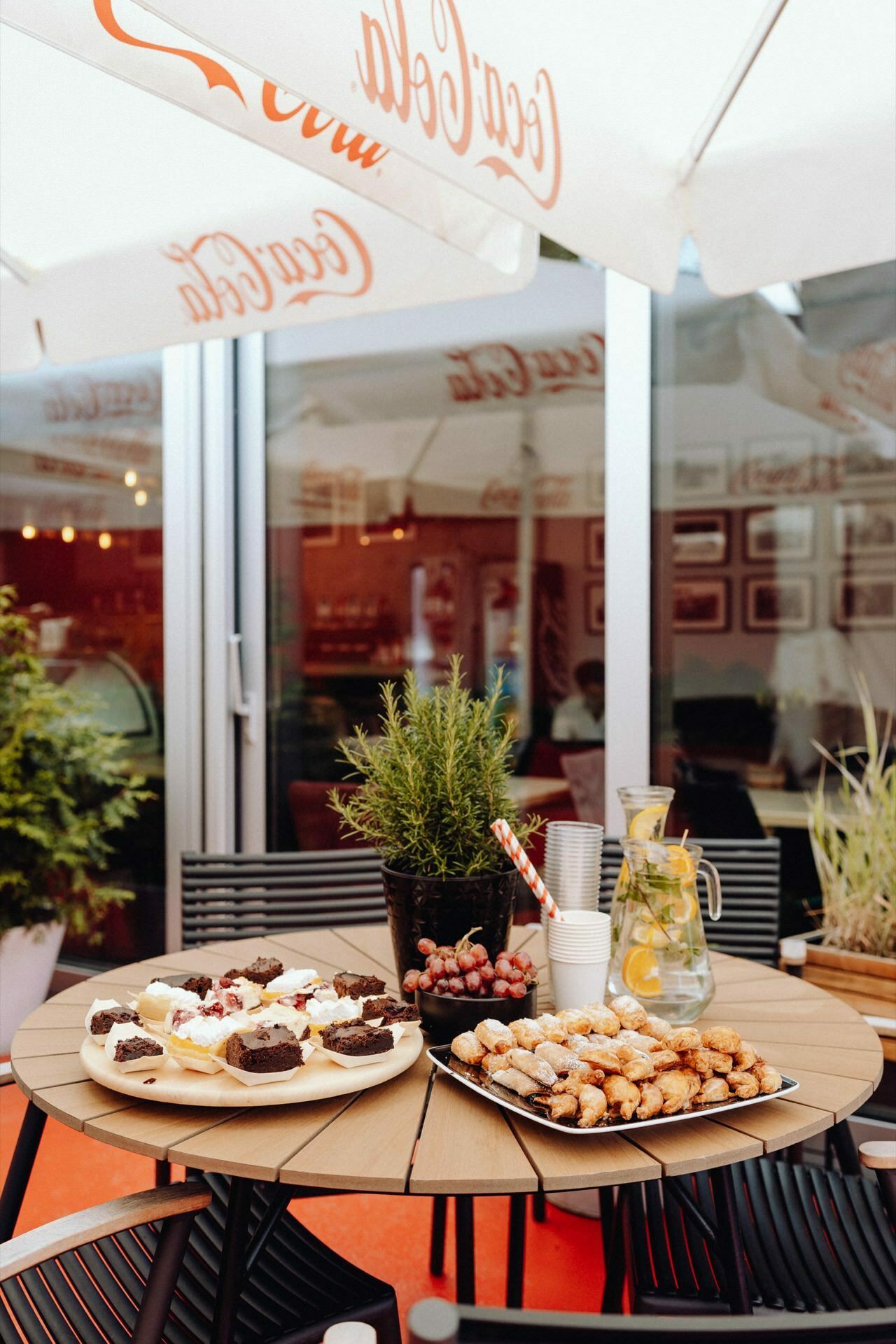 The outdoor cafe features a table with a variety of cakes, grapes and a pitcher of lemon water. The scene reminiscent of an event photo-op is enhanced by a large Coca-Cola umbrella shading black metal chairs. Potted plants and a store window complete this picturesque snapshot.  