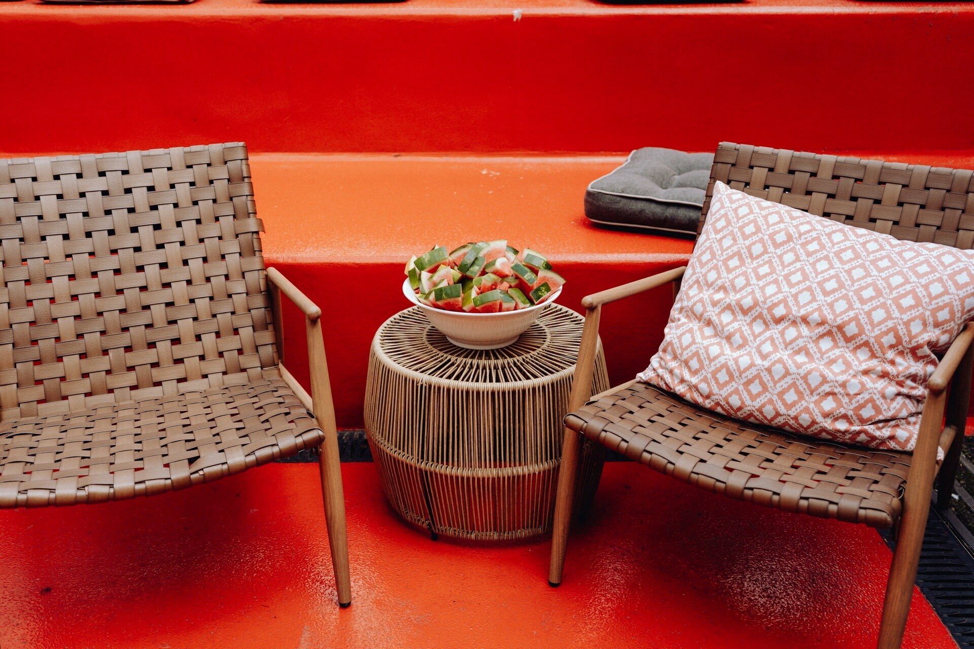 Two woven chairs with brown seats and wooden legs stand on a bright red surface. Between them stands a small round wicker table on which stands a bowl filled with sliced watermelon. In the background, a gray cushion rests on one of the steps, capturing the essence of event photographer Warsaw.  