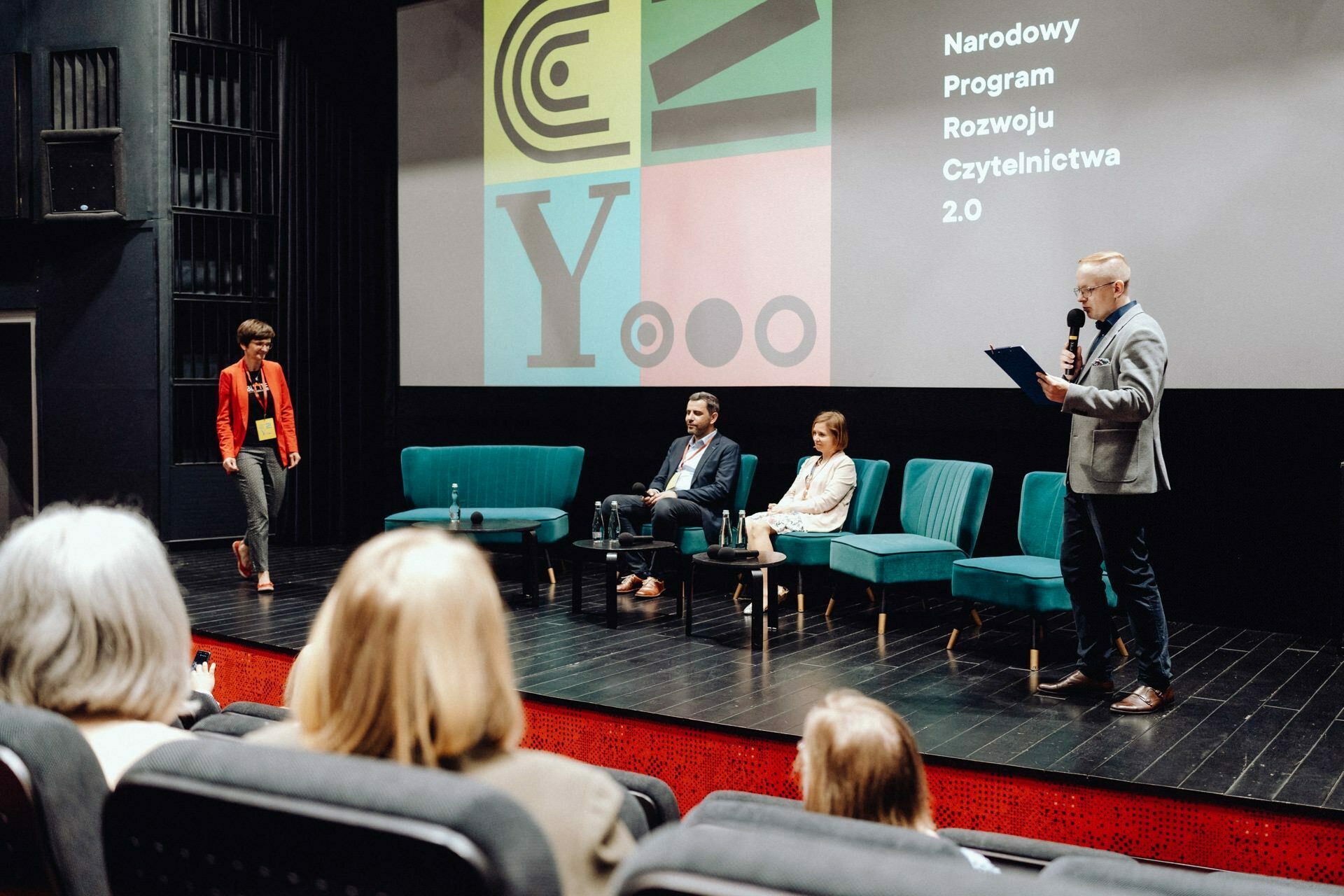 A group of four people are on stage in a conference room. One person, perhaps a photographer from Warsaw, stands on the right with a microphone and a pad, while another approaches them from the left. Two people sit in the middle on a couch, while an attentive audience sits in front.  