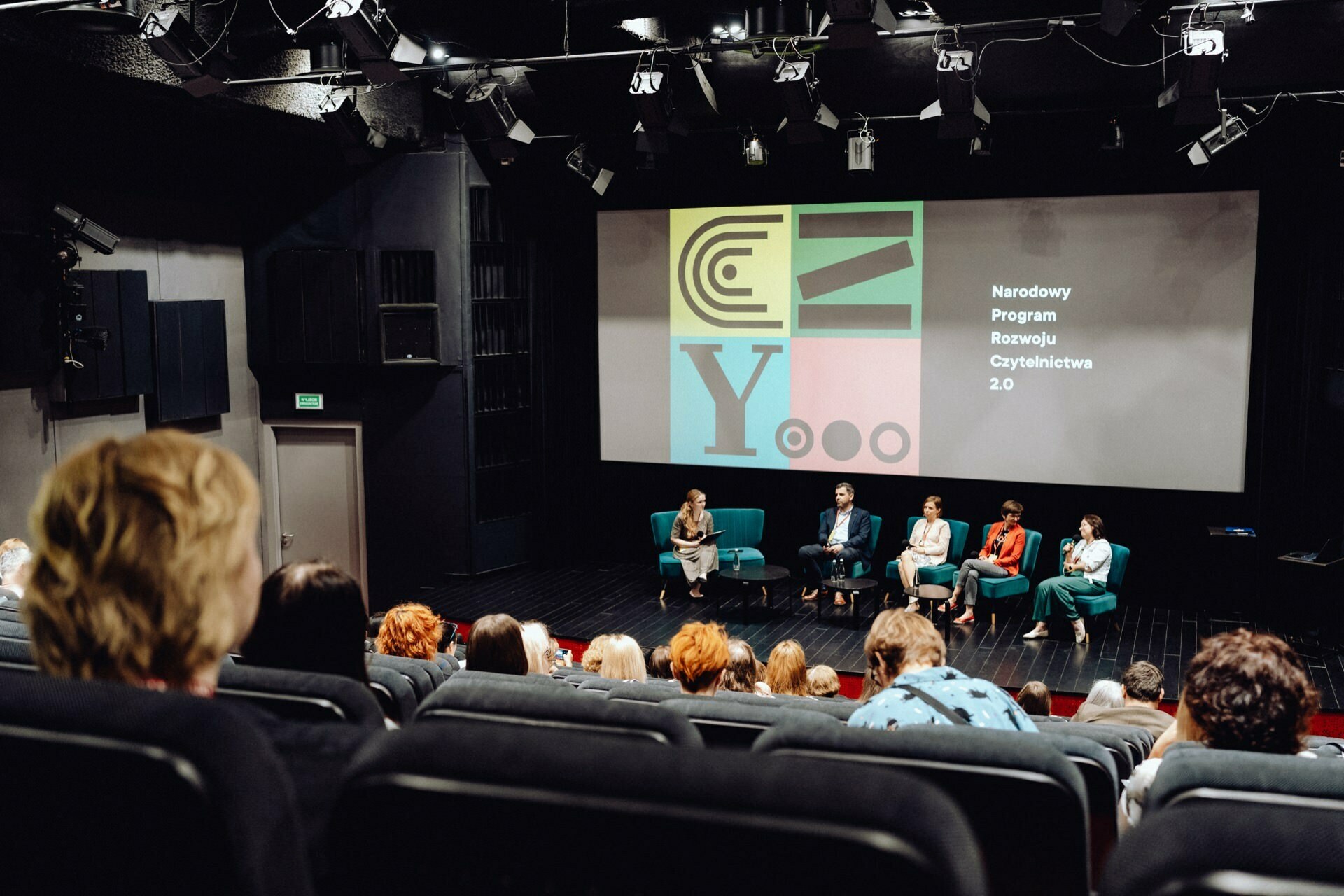 A group of six people sit on chairs on a stage during a panel discussion in a dimly lit auditorium. Behind them, a screen displays graphics and Polish text with the words "National Reading Development Program 2.0." An event photographer from Warsaw captured the engaged audience members sitting in the foreground.  