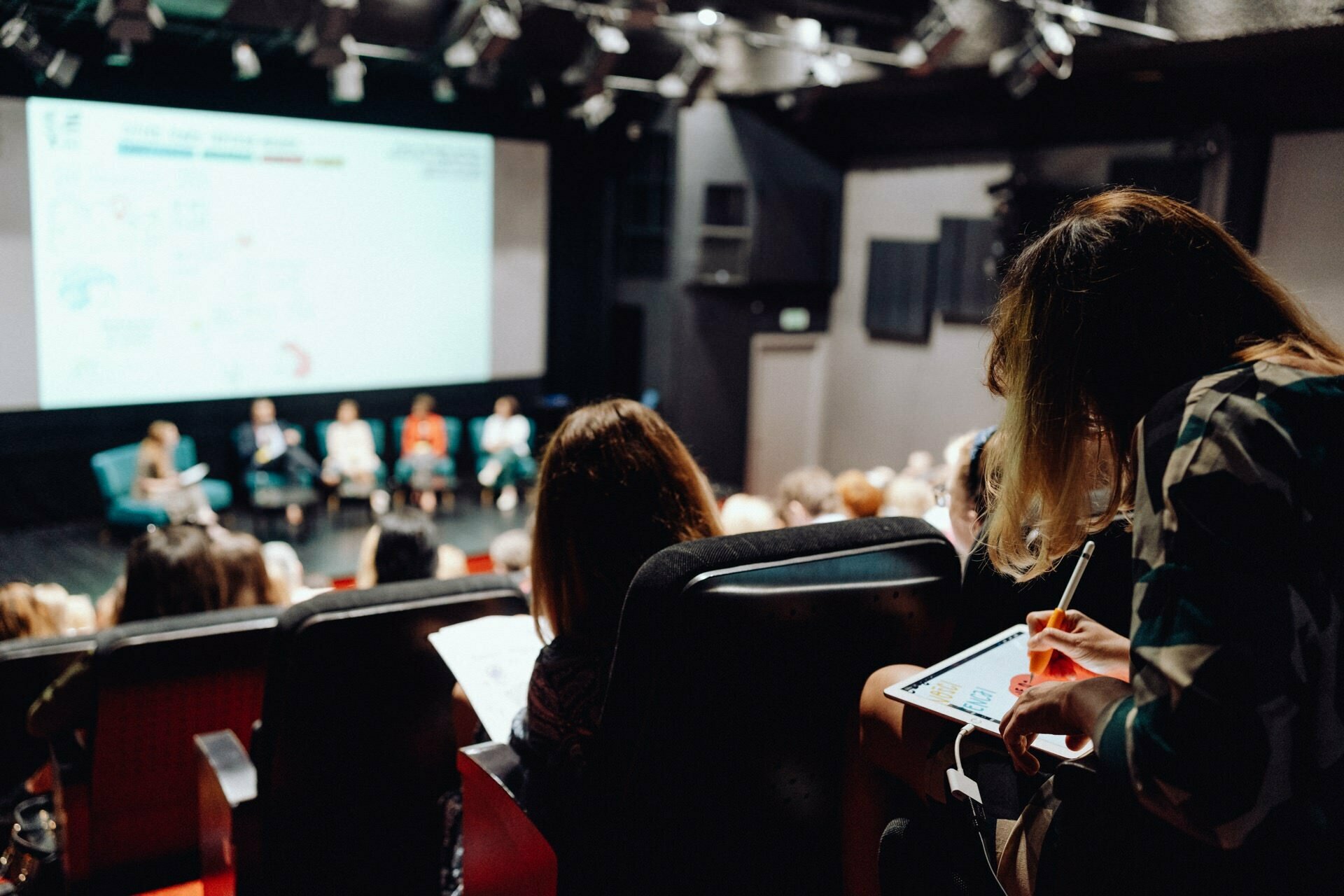 A large group of people are sitting in an auditorium facing the stage, where five people are sitting on a panel with a presentation screen behind them. The center of attention is a woman in the foreground taking notes on a tablet or notepad, captured as part of a photo essay of the event by a photographer for the event. 