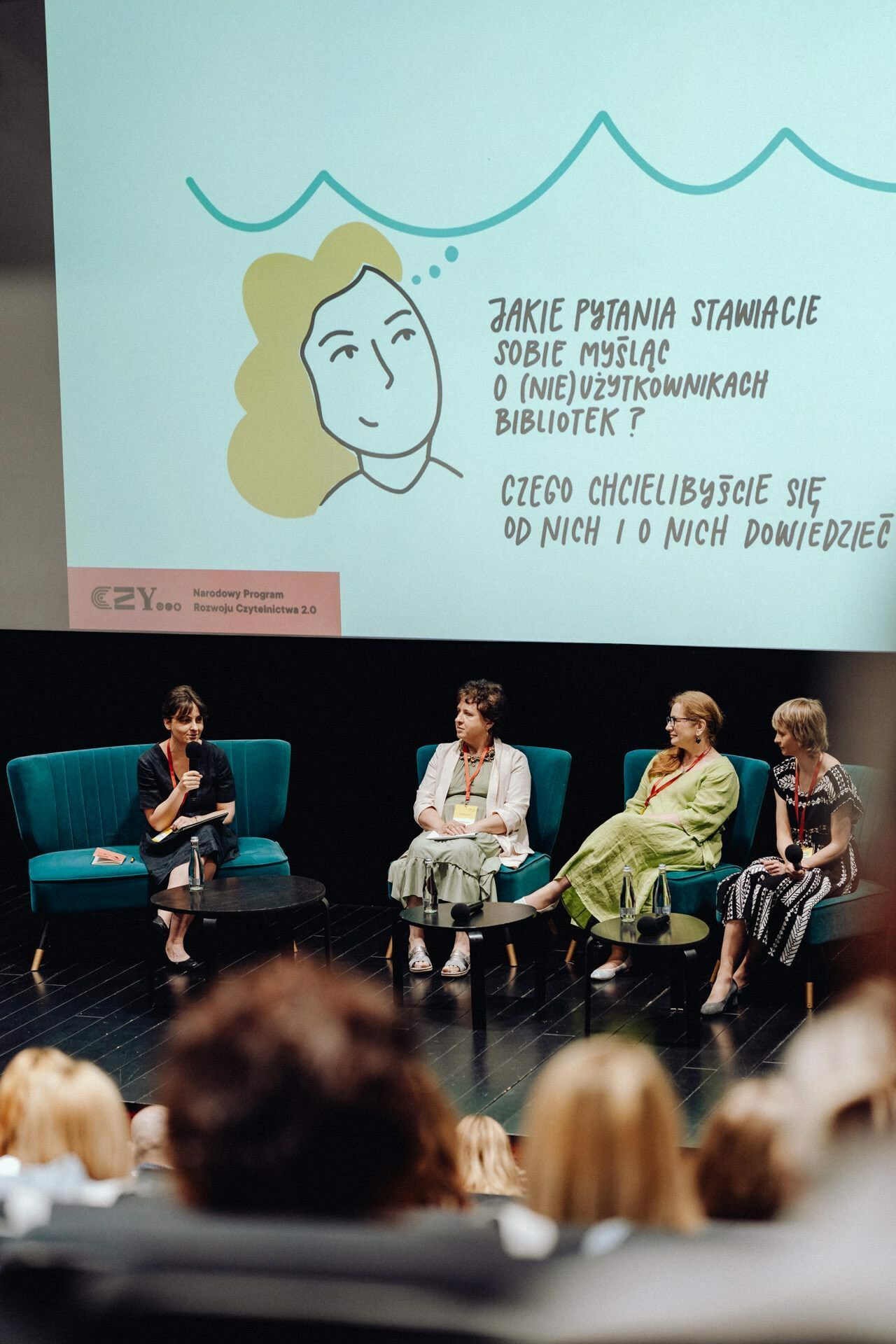 A panel discussion with four women sitting on stage, each with a microphone. They are engaged in conversation. Behind them is a large screen displaying an illustration and text in Polish. A photographer for the event from Warsaw captured this lively interaction. The audience is partially visible in the foreground.    