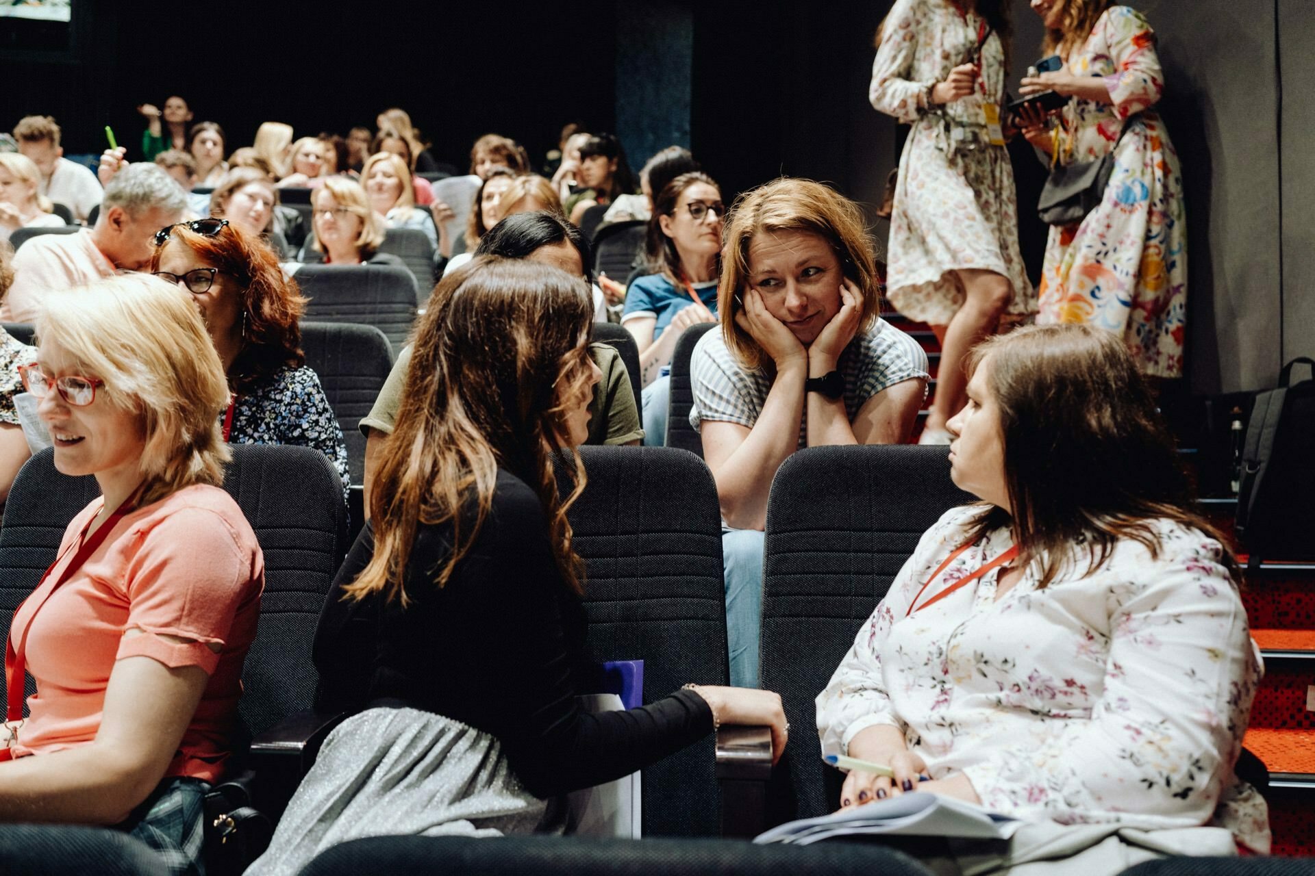 A group of people are sitting in an auditorium having a conversation before the event begins. We focus on two women in the foreground having a lively discussion, while an event photographer Warsaw captures other people in the background who are also talking and preparing for the event. 