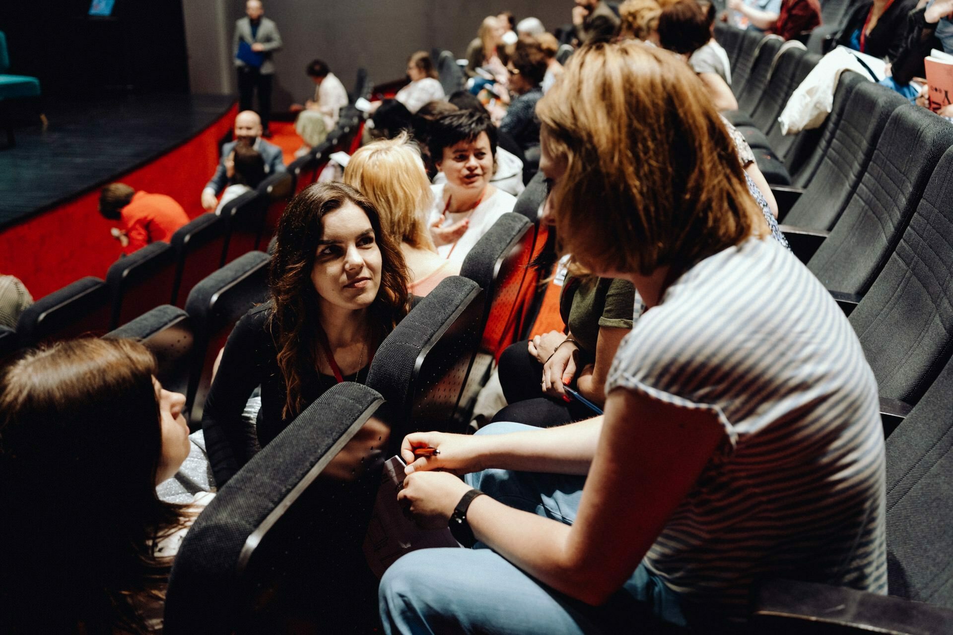 A group of people sitting in theater seats are having a conversation while waiting for the show to start. They are casually dressed and look engaged. More audience members can be seen in the background, indicating a lively atmosphere in the audience, perfectly captured by a photographer from Warsaw.  