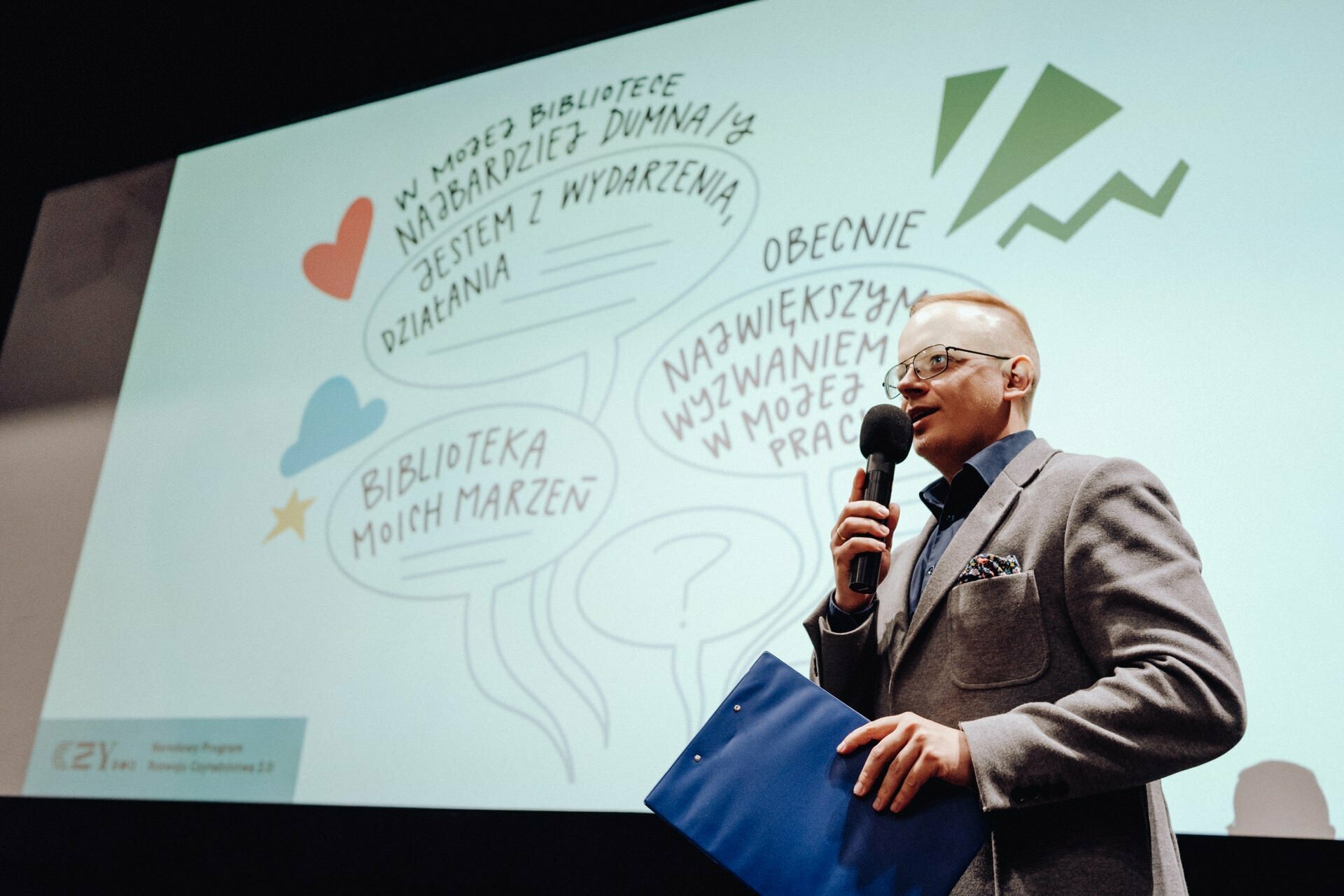 A person wearing glasses and a suit holds a microphone and a blue briefcase and speaks in front of a screen displaying balloons with text in Polish. In the background are colorful balloons with various phrases and decorative elements, beautifully captured by a photographer from Warsaw documenting the event. 