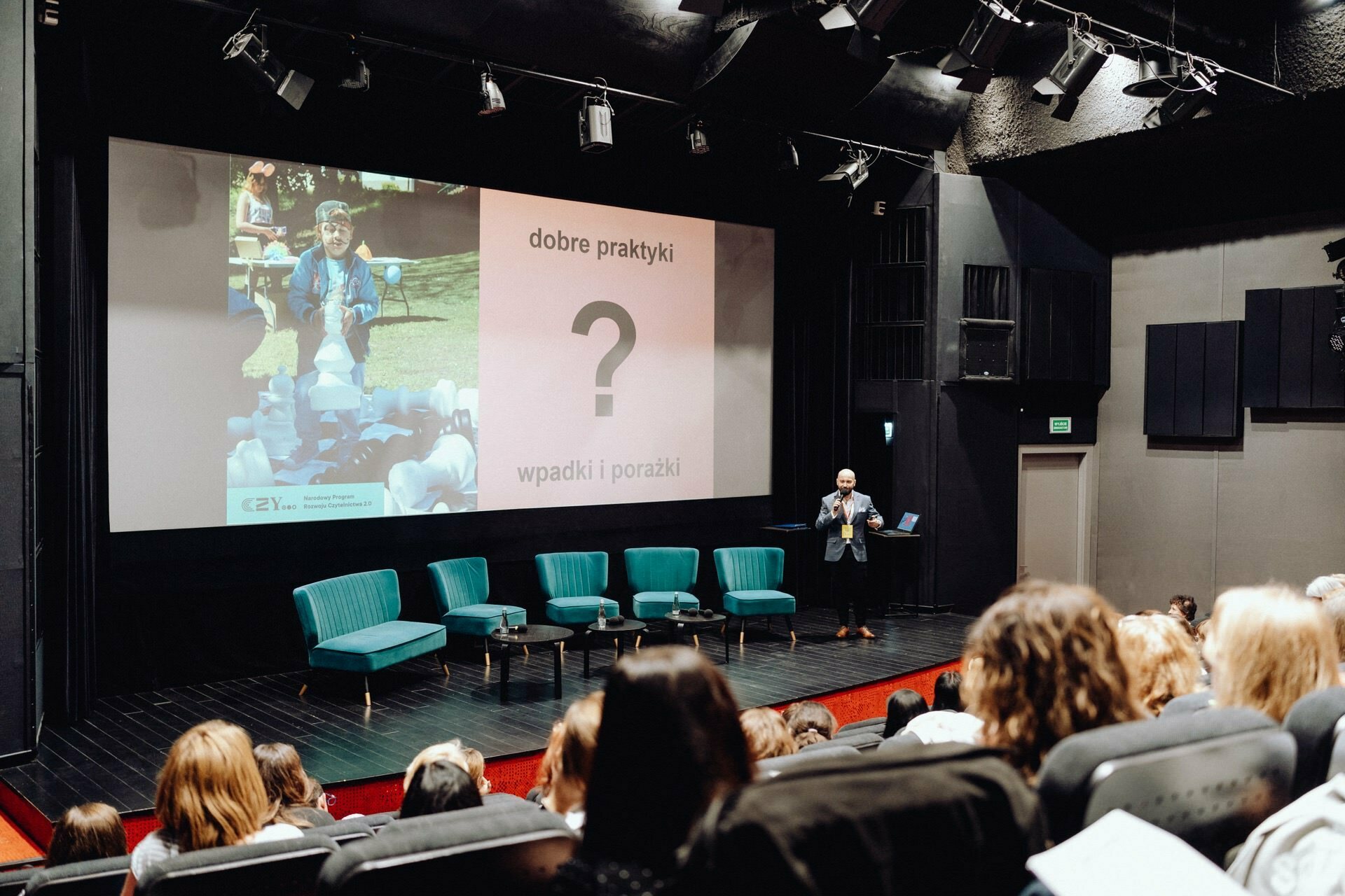 The presenter stands on a stage in a theater room and speaks to the audience. At the back, a large screen displays a slide with text and a photo by a *photographer from Warsaw* showing a person conducting outdoor activities with children. There are several turquoise chairs set up on the stage in the room.  