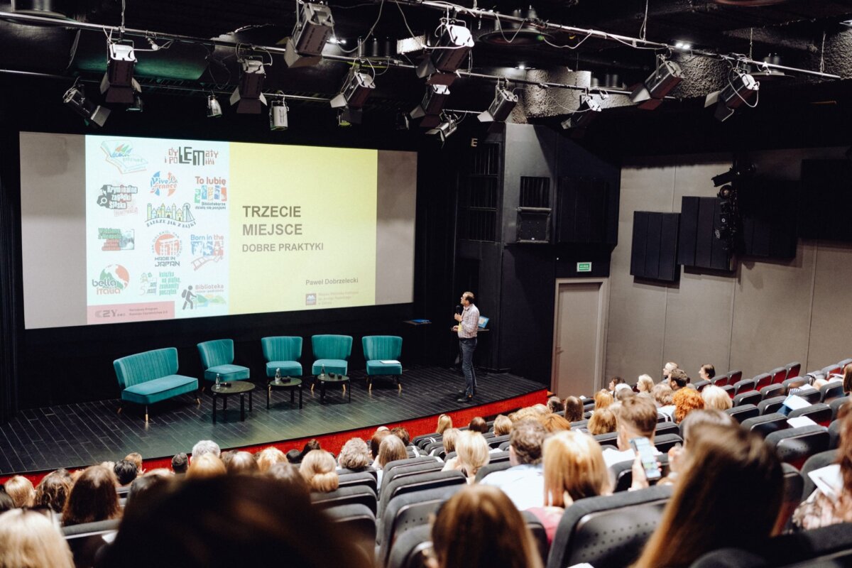 The speaker stands on a stage and performs before an audience in a theater. Behind them, a presentation slide entitled "Third Place Good Practices" is displayed on a large screen. Several empty turquoise chairs were placed on the stage. The audience is seated and focused, as captured by the event photographer warszawa.