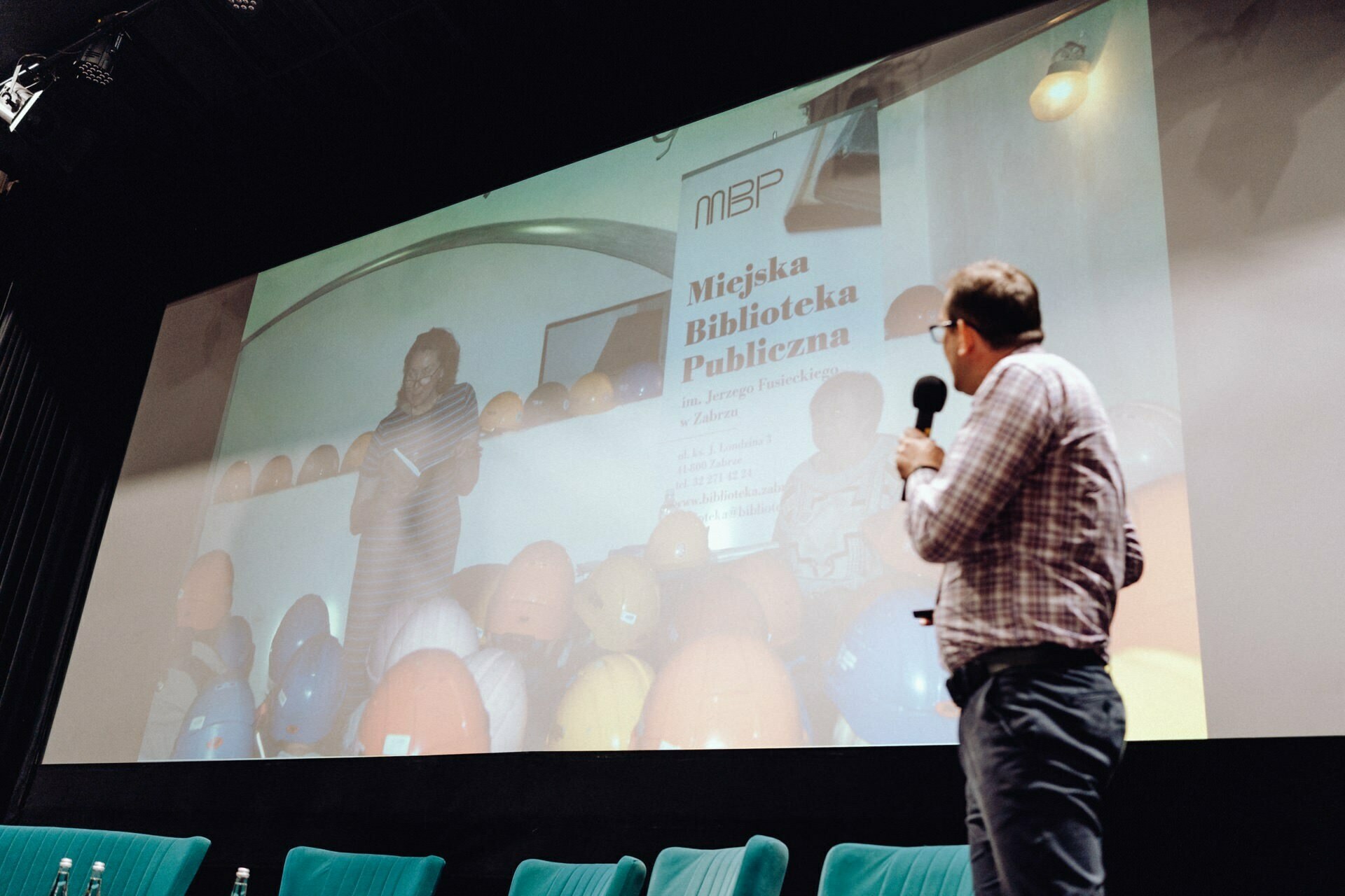 The presenter stands on the stage with a microphone, facing a large screen that shows an image of the speaker with a banner reading "Municipal Public Library." There are several turquoise-colored chairs on the stage, capturing the essence of the lively conference. The photo coverage of the event was expertly taken by a photographer from Warsaw.  