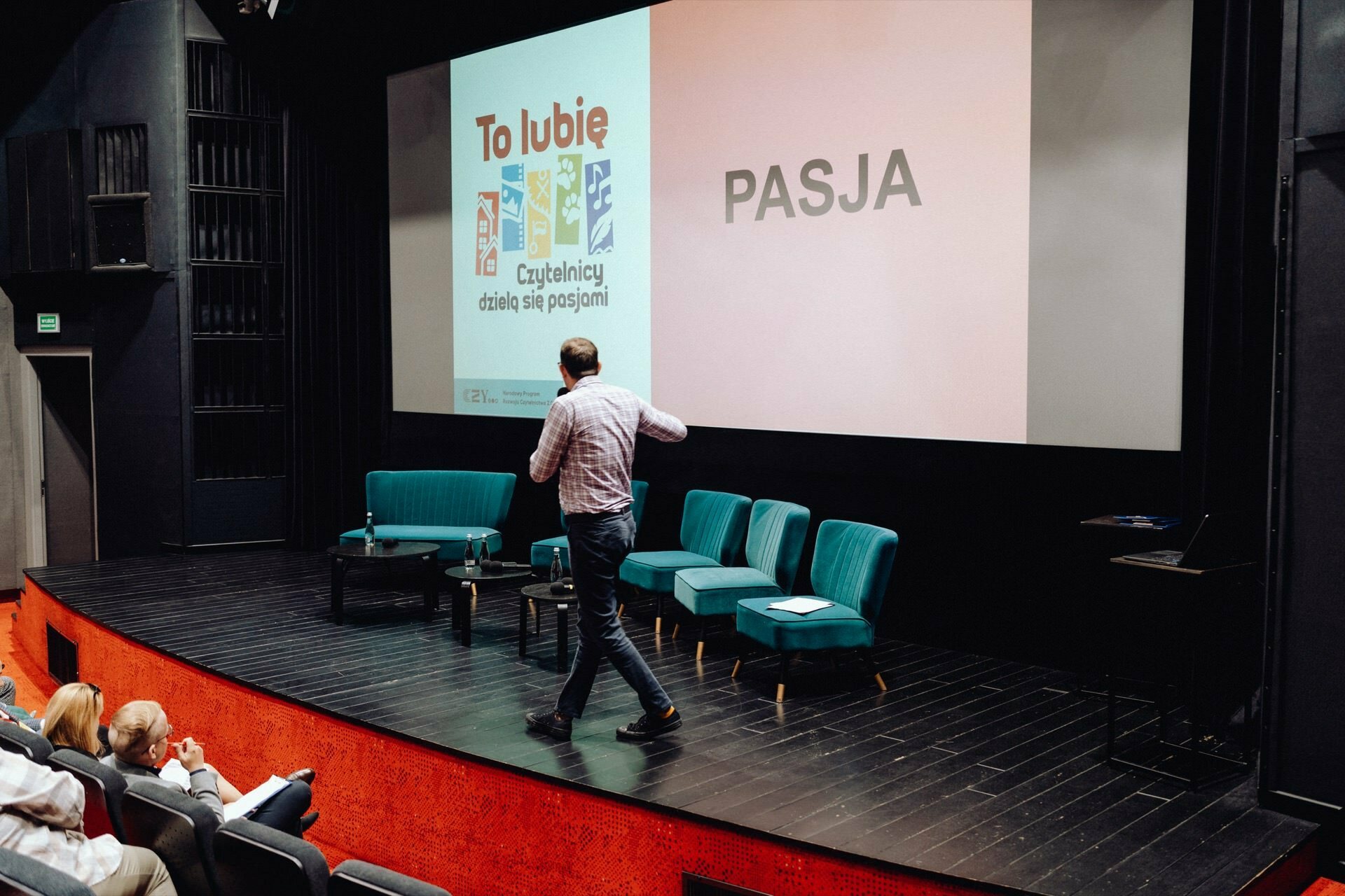 The man stands on stage in front of the audience and points to a large screen, on which the word "PASSION" and a colorful graphic with the words "This I like" appear. Four turquoise chairs and a small table are set up behind him. The scene appears to be part of a photo essay of the event captured by a photographer for the event.  