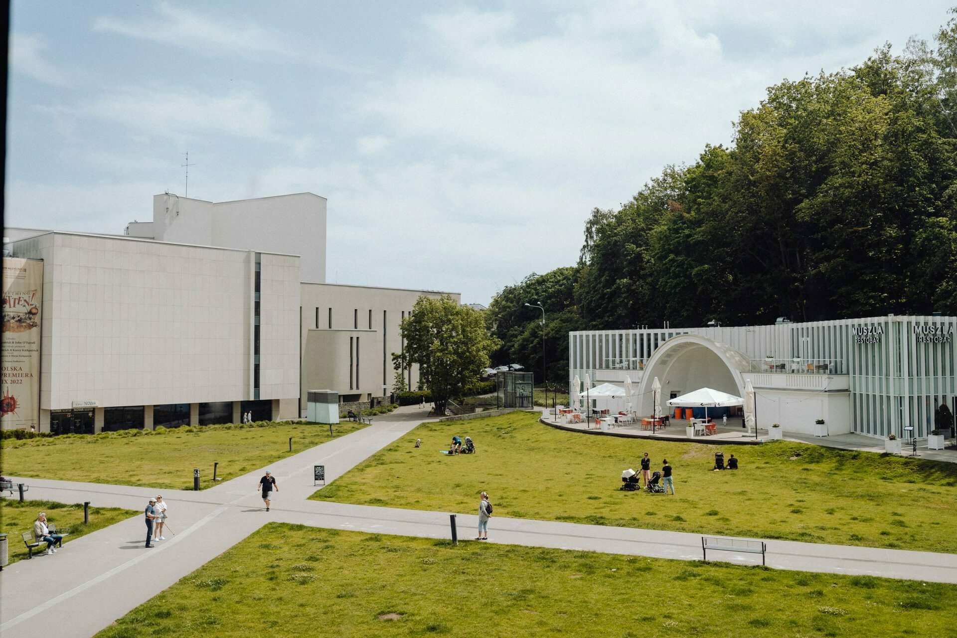 A modern cultural building with a large white facade and a glass arched stage is surrounded by green lawns and scattered trees. People walk, sit and relax on the grass and paths under clear blue skies - a perfect scene for an event photographer Warsaw to capture. 