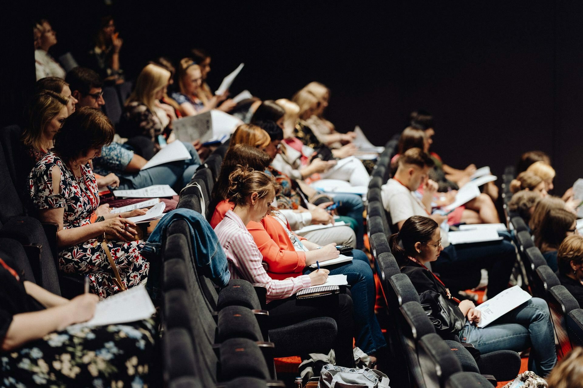 A large group of people sit in tiered rows in the auditorium, carefully reading or taking notes on papers lying on their laps. The audience consists of people of all ages, dressed in casual and semi-formal attire. Event photographer Warsaw captures the scene of this educational or professional meeting.  