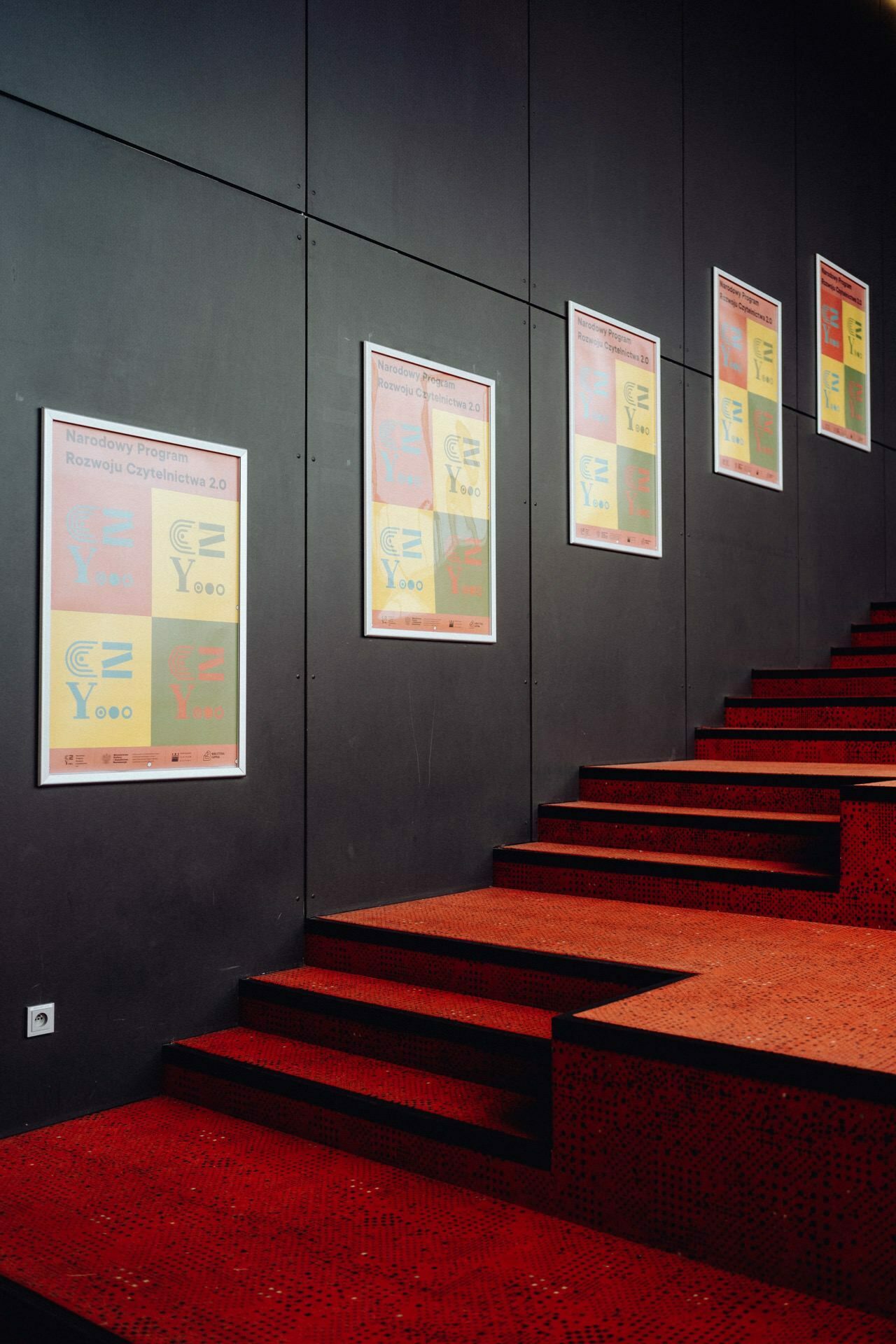 A staircase with red steps and a black wall decorated with six identical posters. The posters, arranged in a diagonal line, feature colorful text and graphics promoting the program. The stage is well-lit, highlighting the vivid colors - ideal for event photojournalism or capturing memories by a Warsaw event photographer.  