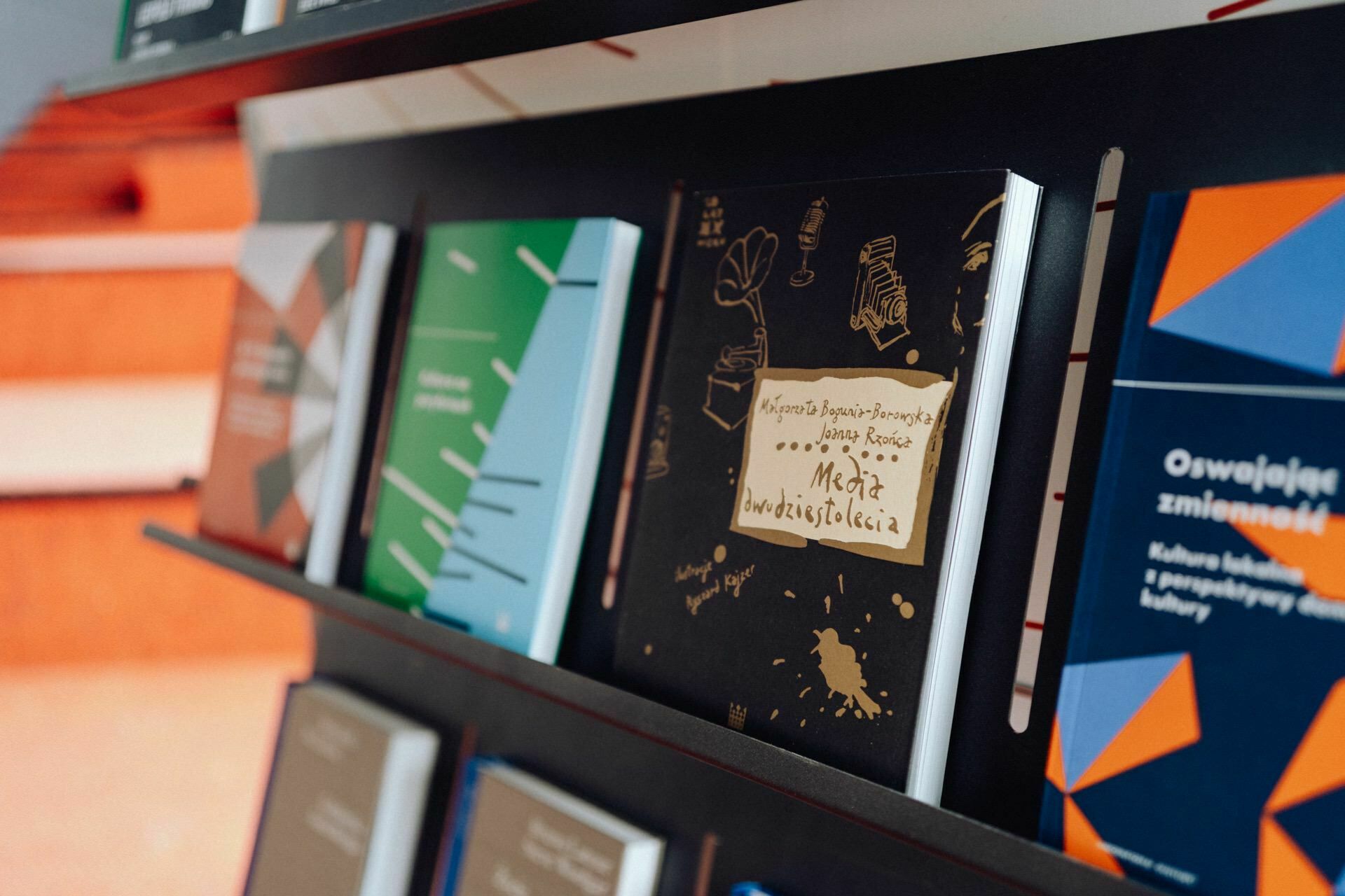 A row of books displayed on diagonal shelves in a bookstore or library. Among them, one prominent book has a dark cover with gold illustrations and handwritten text. Other books have colorful covers with abstract patterns and various titles, resembling captions from a photo report of the event taken by a photographer for the event.  