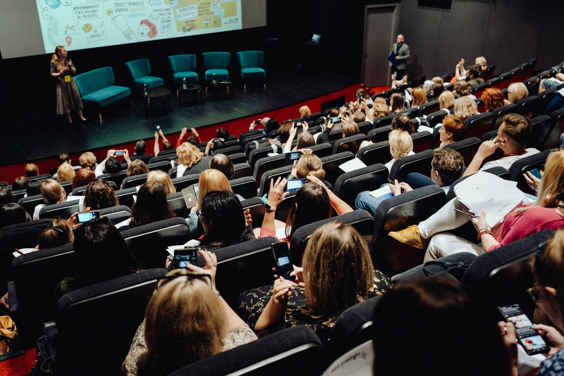 A crowded hall with people sitting in tiered rows, many holding smartphones. Several people take photos or videos of a woman speaking on stage next to a large screen displaying colorful graphics and text. There are four empty chairs on the stage, perfect for a comprehensive photo essay of the event.  