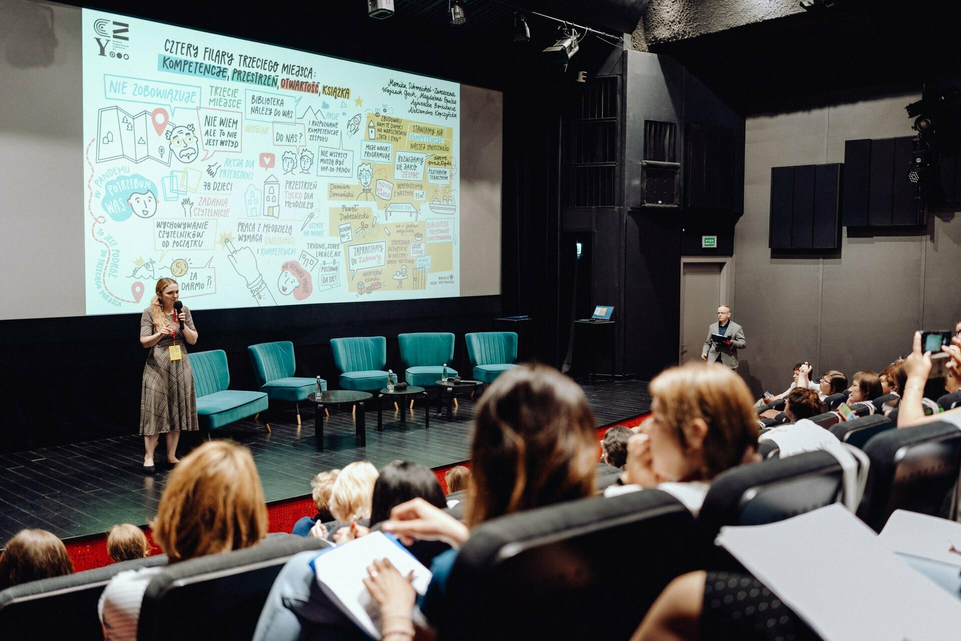 A woman stands on stage next to a large illustrated presentation screen in a conference room with theater-style seating. The audience sits and takes notes while a man near the stage points to the screen. Several blue chairs stand on the stage, captured perfectly by event photographer Warsaw.  