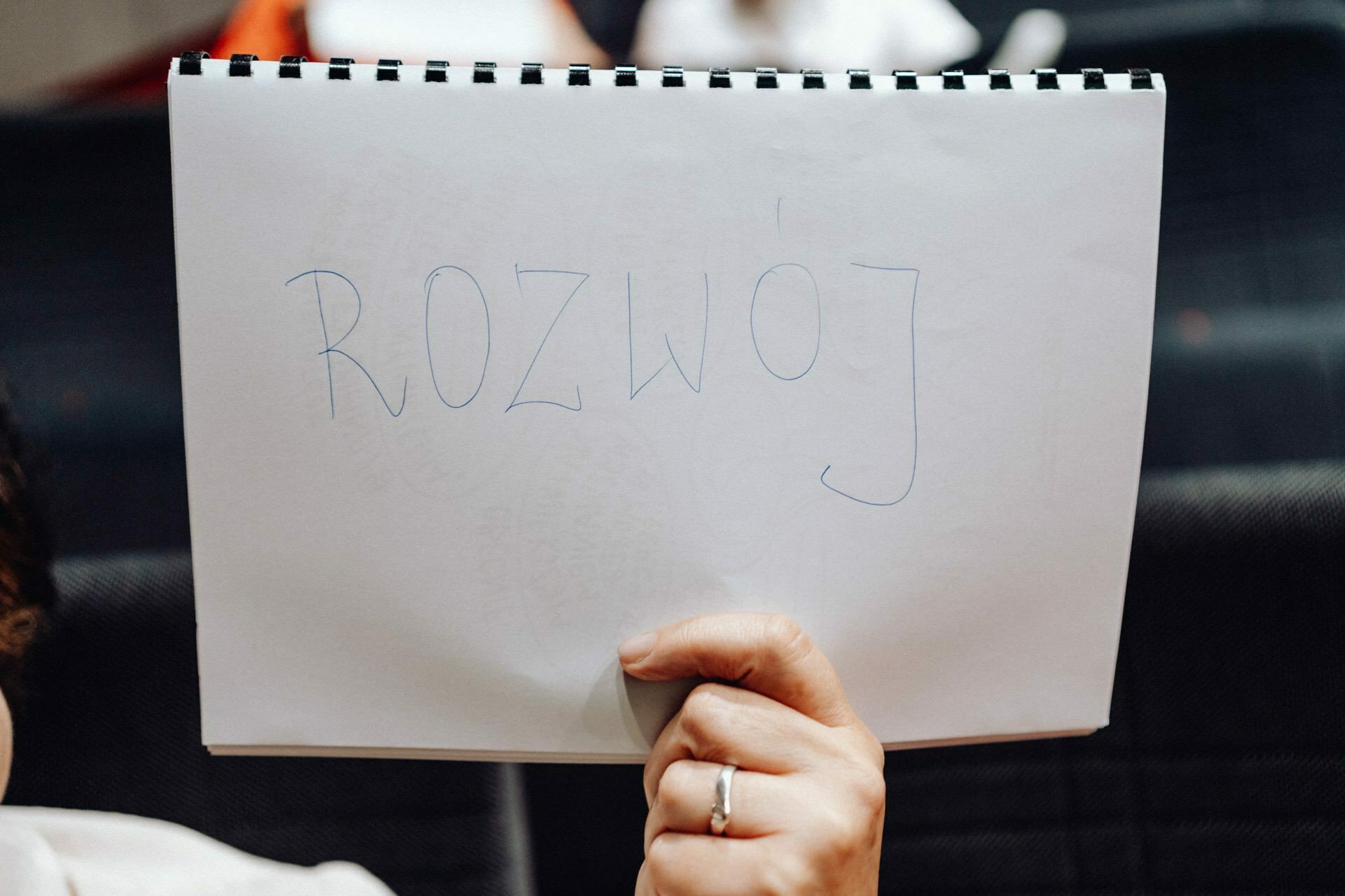 The person is holding a white notebook with the word "DEVELOPMENT" written in blue ink. Partially visible is the hand of the person with the ring, holding the bottom of the notebook. The background, blurred and dark, suggests a room setting - perhaps captured by the photographer at the event.  