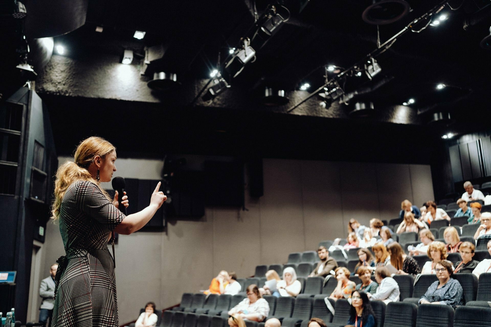 A person stands on a stage, holds a microphone and speaks to an audience seated in a dimly lit auditorium. The audience is focused on the speaker, with some taking notes and others listening intently. The room has a modern decor with black walls and overhead lighting, which was perfectly captured by our photographer at the event.  