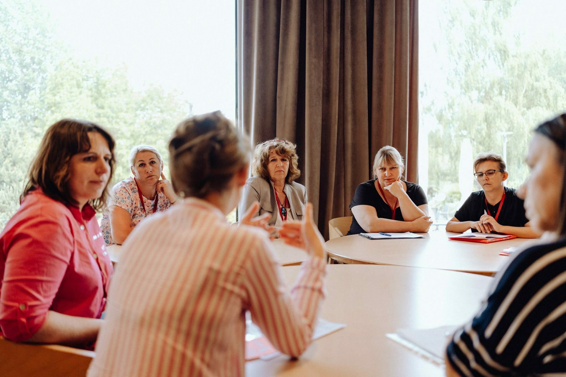 Six people sit at a table immersed in a serious discussion. The person in the foreground, with his back turned to the camera, gestures with his hands as he speaks. Light from the large windows illuminates the room, behind curtains partially drawn to the side, which was perfectly captured by the photographer for the Warsaw event.  