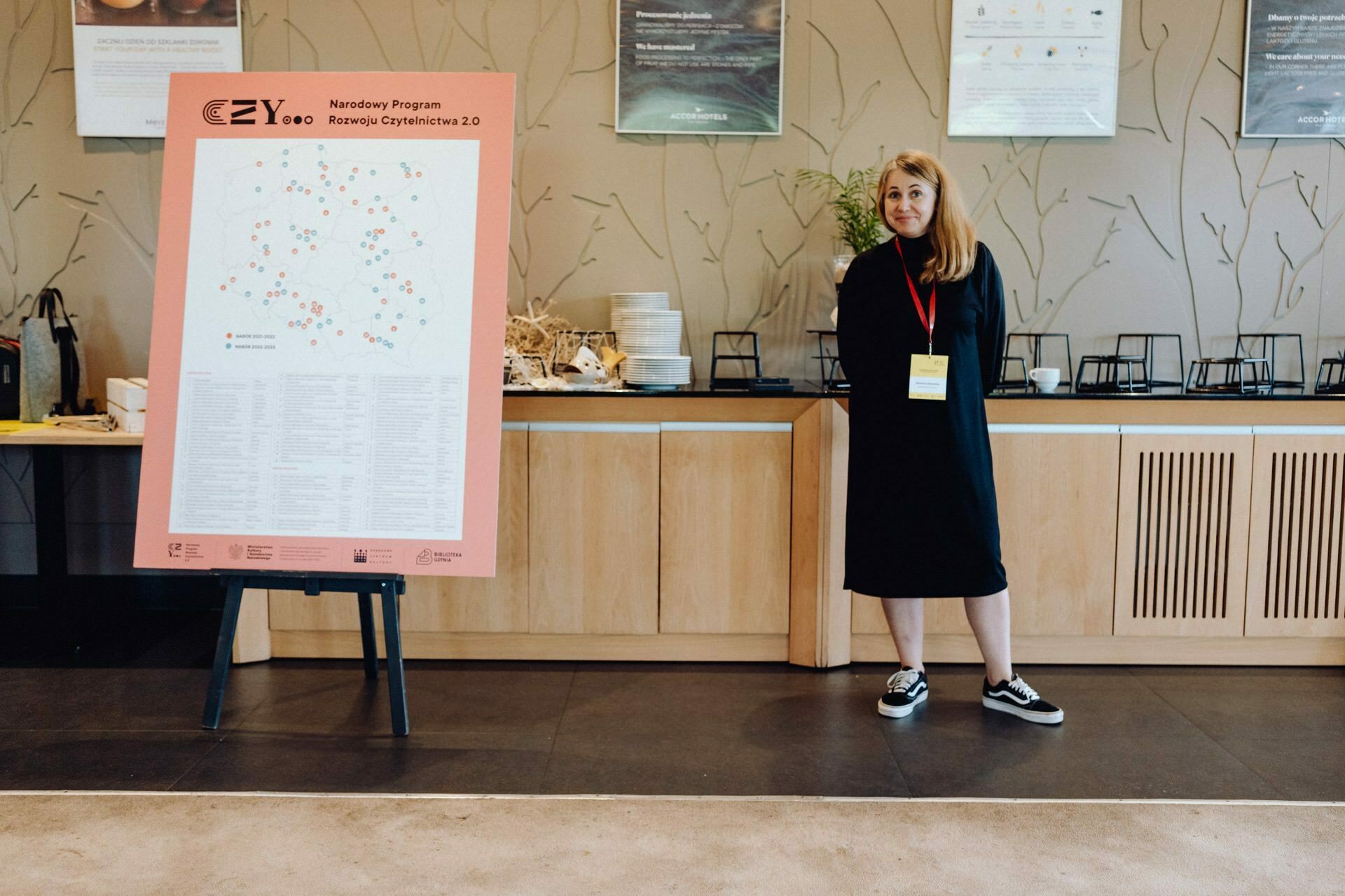 A woman is standing next to an easel, holding a large poster with a map and text. She is dressed in a black outfit with a red lanyard and is smiling slightly. Behind her is a counter with decorations and framed photos on the wall, all taken by a talented event photographer from Warsaw.  