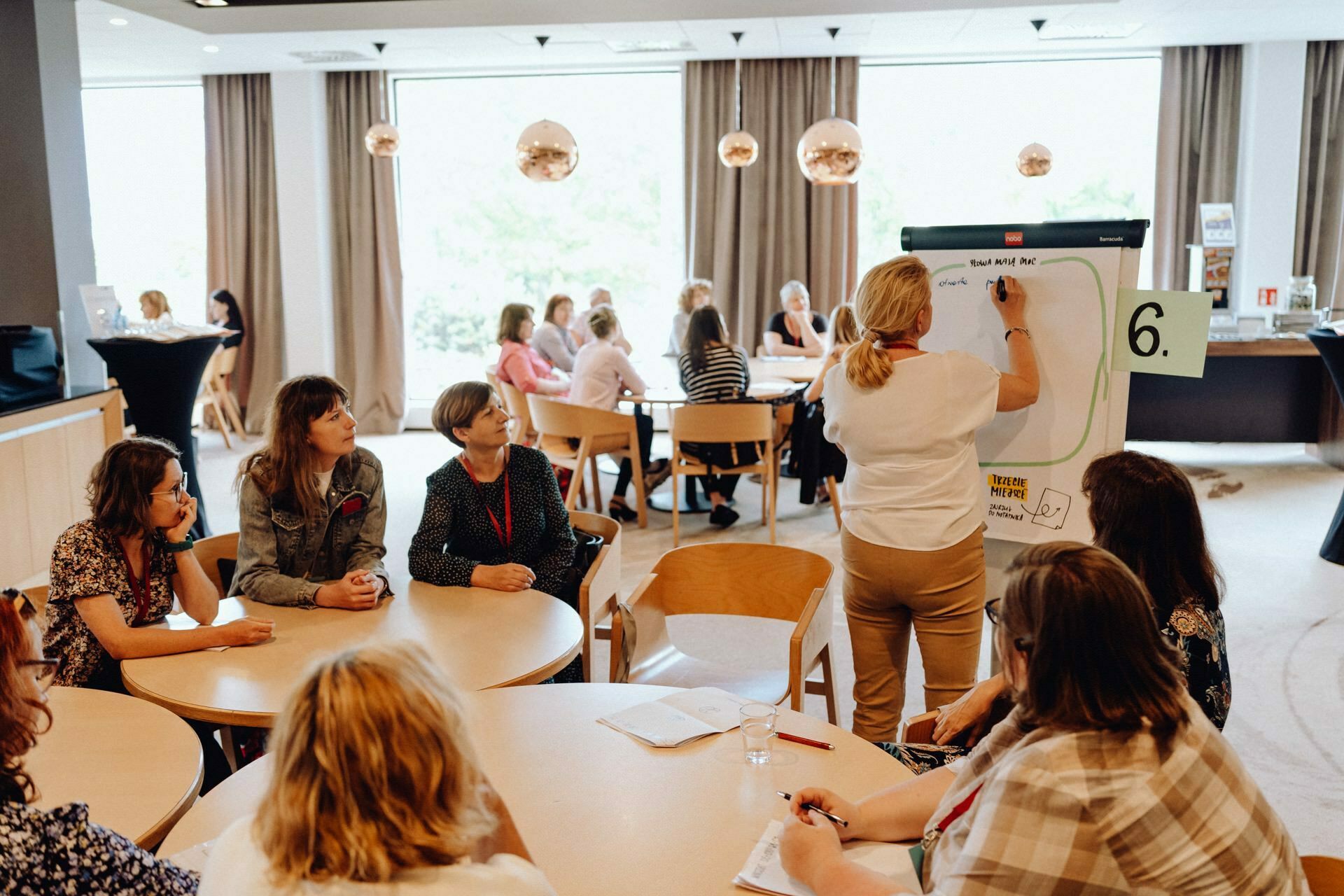 A group of people gather in a well-lit room with many round tables. A woman stands at an easel with a large notebook and writes, while others sit around the tables and watch intently. The surroundings suggest a communal workshop or meeting, perfect for an event photo essay by a photographer from Warsaw.  