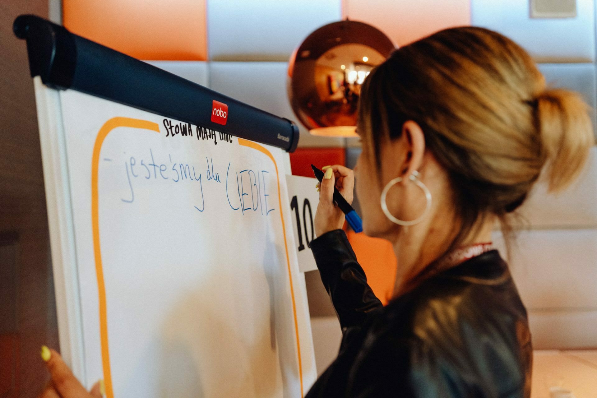 A person with his hair tied in a ponytail, wearing a brown leather jacket and hoop earrings is writing on a blackboard. The board already has text written in a language other than English. In the background of the event photo report is a reflective copper ball and abstract wall decor.  