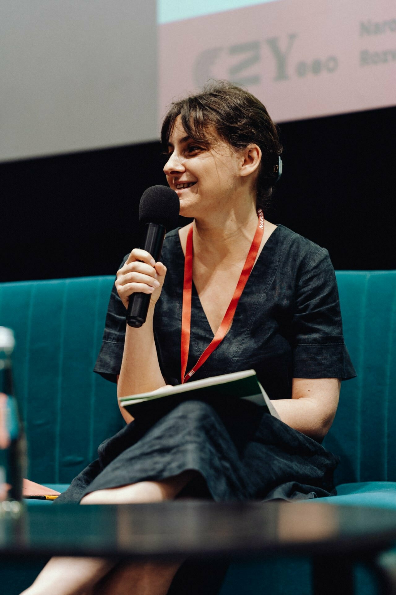 A person with dark hair, wearing a dark dress and a red lanyard, sits on a turquoise couch, speaks into a microphone and holds a notepad. They are smiling and look as if they are participating in a panel discussion. This moment of the event can be perfectly captured by any skilled event photographer in Warsaw.  