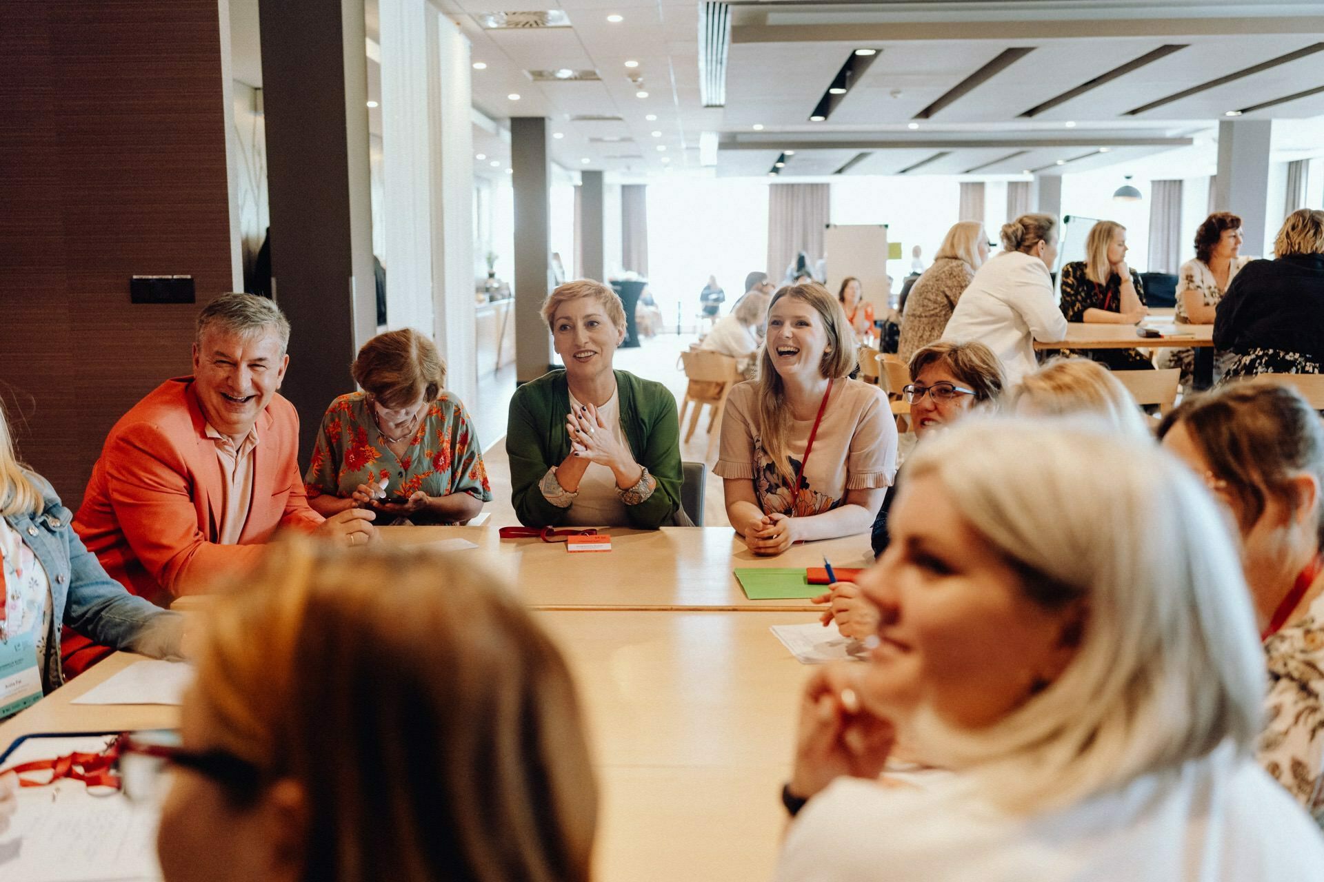 A diverse group of people, seated around a table, engage in lively conversation and laughter during a meeting or workshop in a spacious, well-lit room. This photo report of the event beautifully captures the collaborative and friendly atmosphere. 