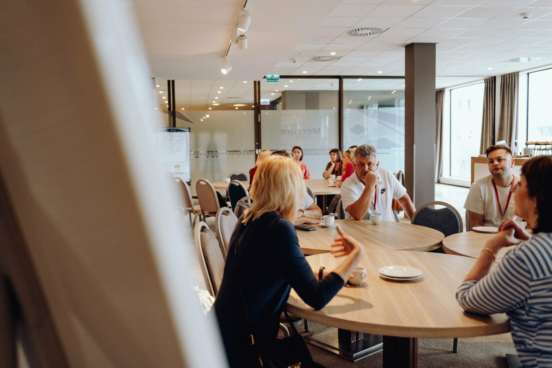 A group of people sitting around tables in a well-lit room, immersed in conversation. The woman in the foreground is gesturing with her hand, which was perfectly captured by a photographer from Warsaw. The room has large windows and modern decor in neutral colors. Plates and cups stand on the tables.   