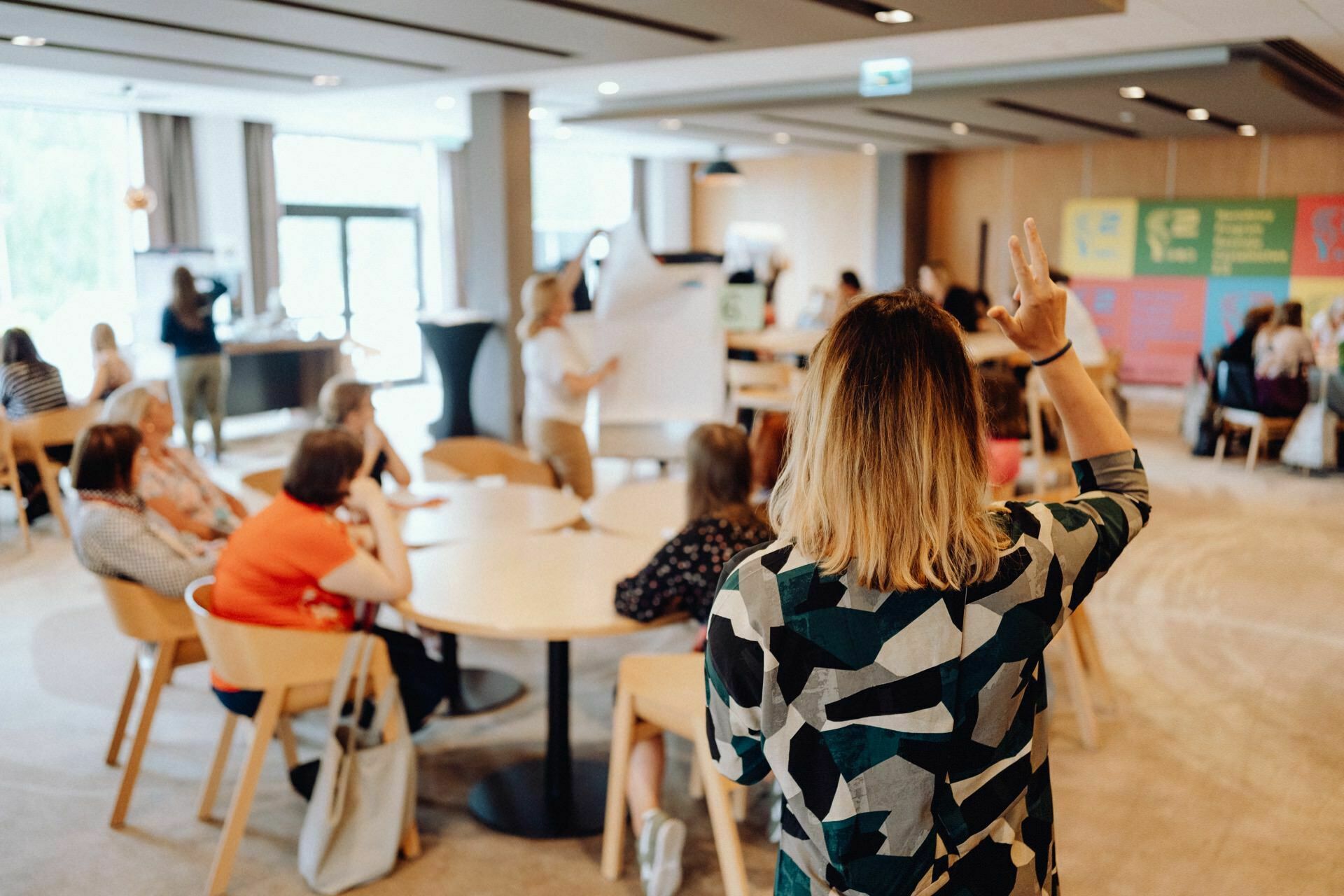 A person with shoulder-length hair and wearing a patterned shirt raises his hand, looking at a group of people seated at round tables. A presentation is made at the front of the room, during which the person points to a large board, beautifully captured by the photographer for the event. 