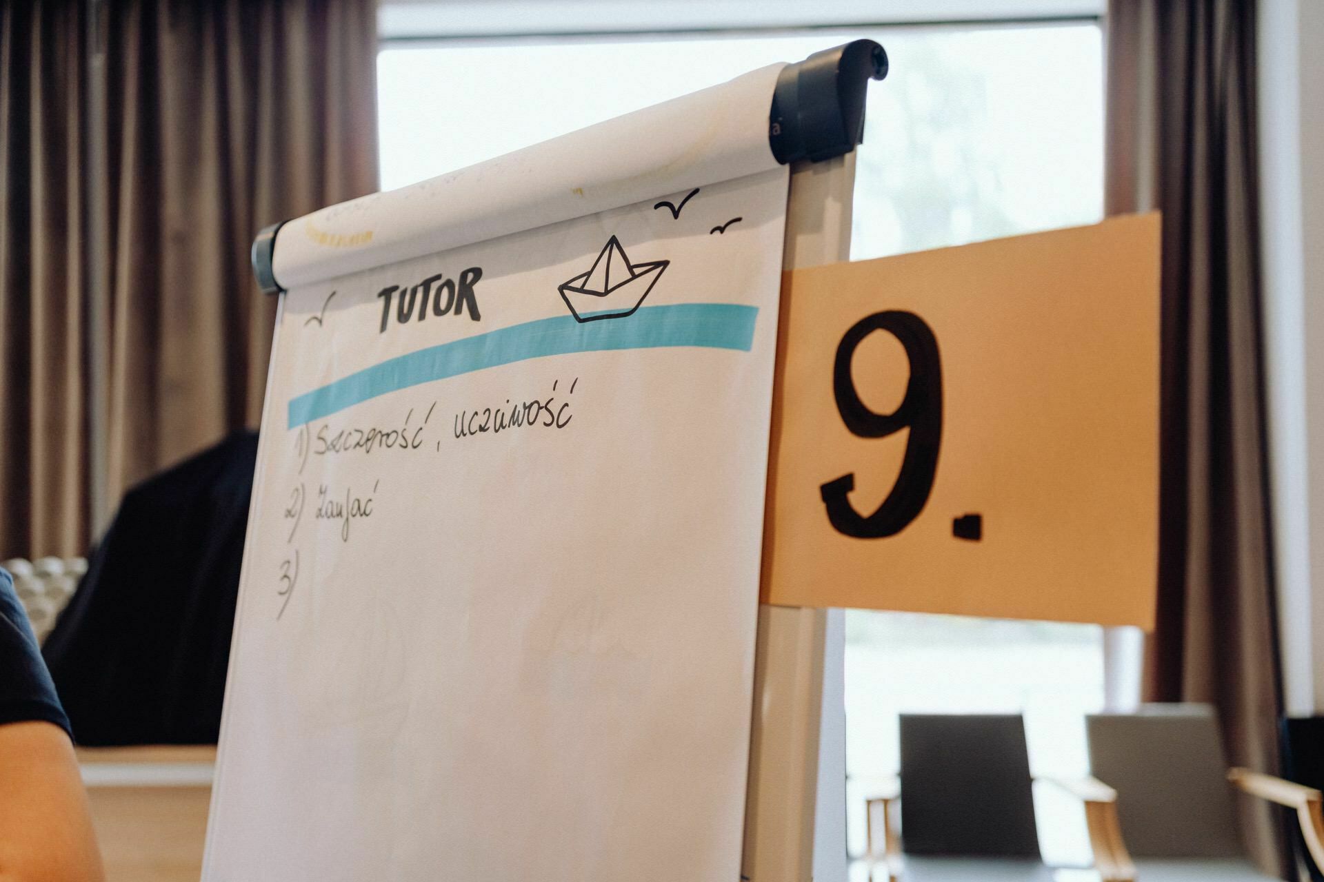 A flipchart stands in the room next to the large card with the number 9. On the flipchart marked "TUTOR" and decorated with an illustration of a paper boat are notes in a foreign language. In the background are curtains and fuzzy chairs, beautifully captured by a photographer from Warsaw during the event.  