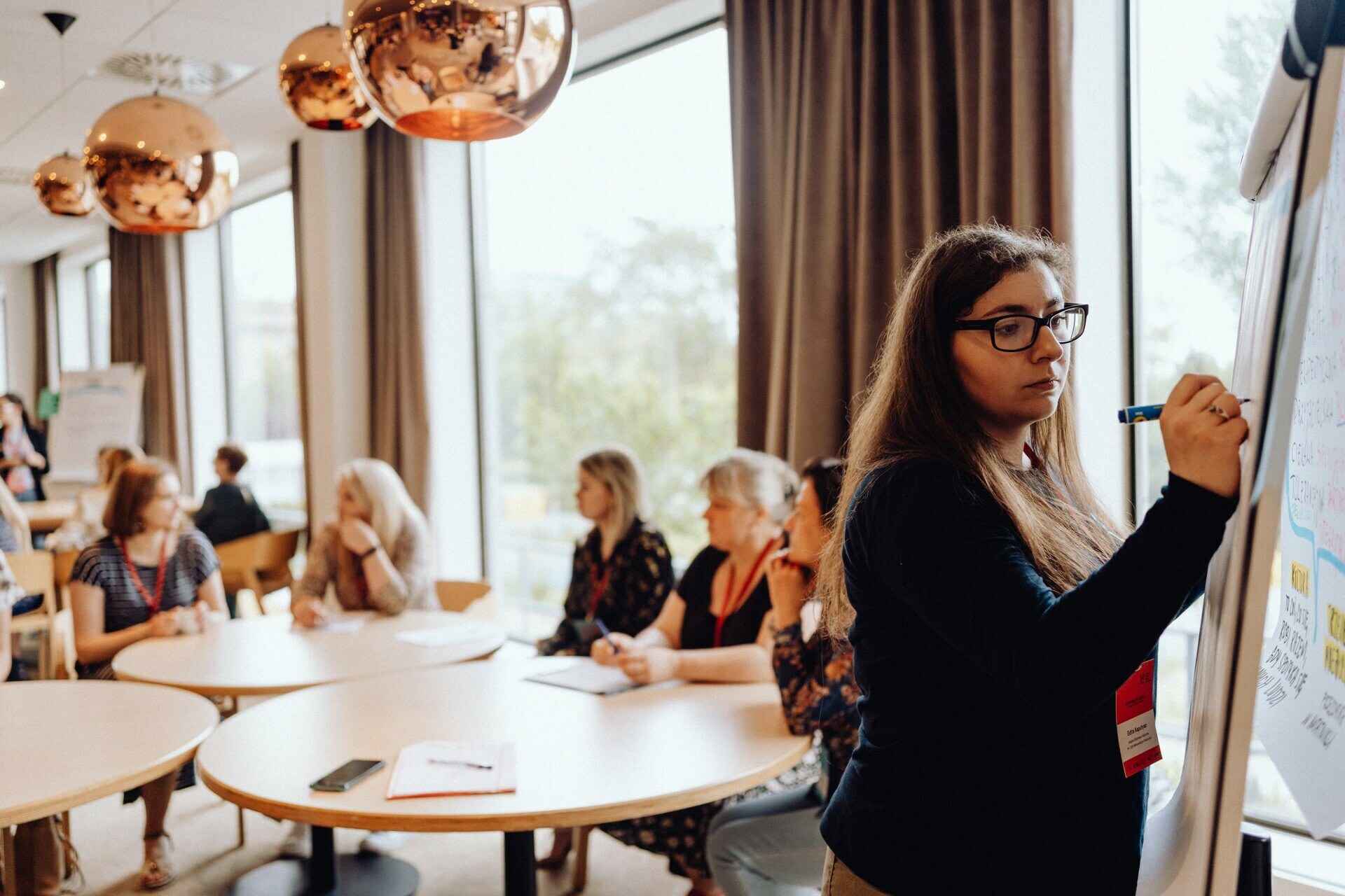 A woman is writing on a flipchart in a bright room with large windows. Several people sit at round tables and watch her intently. The space is ideal for photo coverage of the event, decorated with pendant lamps and there is a warm, professional atmosphere.  