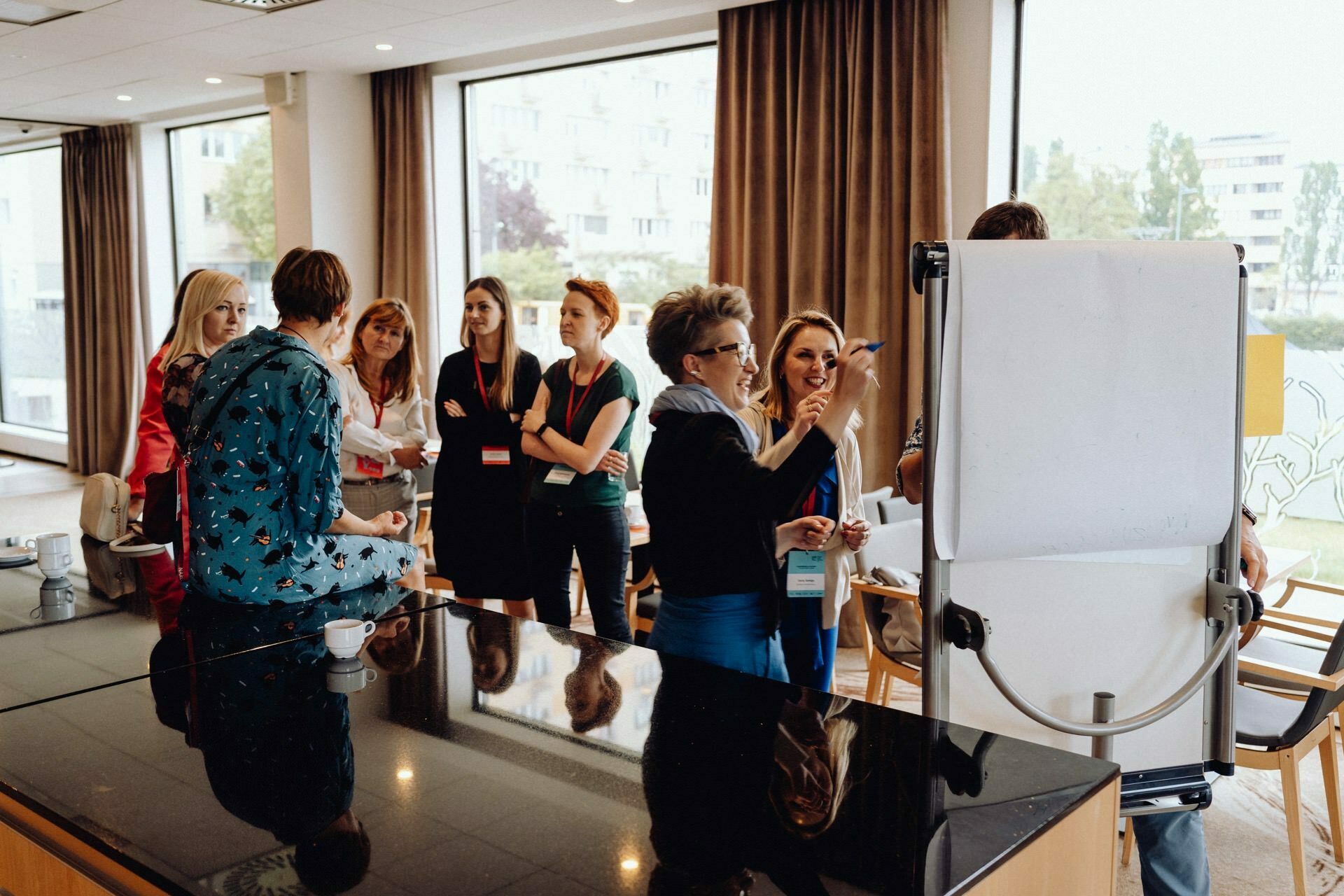 A group of people stand around a flipchart in a conference room. One person writes on the flipchart while the others watch and discuss. The room captured by event photographer Warsaw has large windows overlooking the building and trees outside. In the foreground is a tabletop with a teacup.   