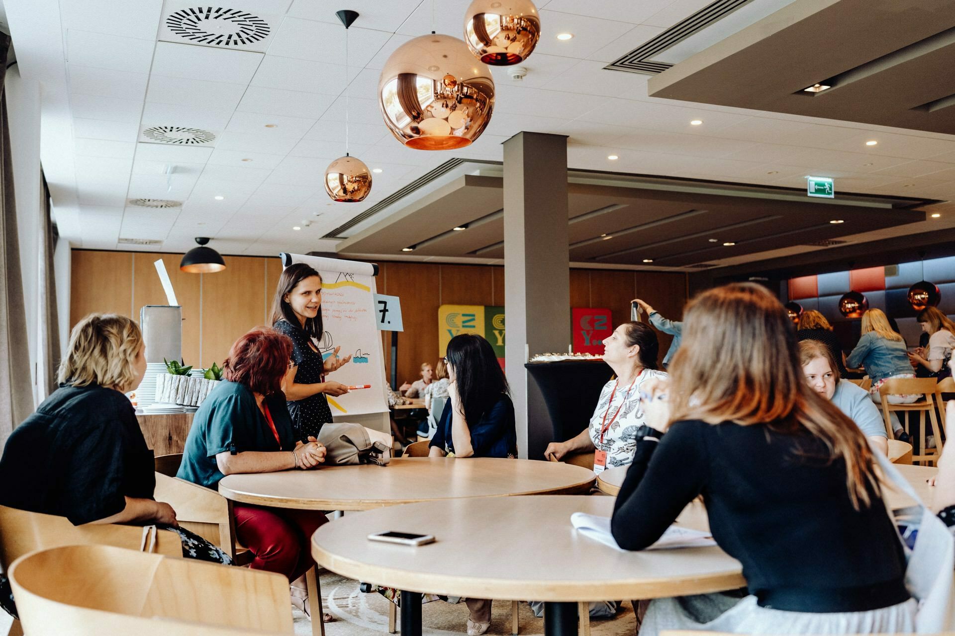 A group of women sit around round tables in a modern, well-lit conference room with wooden accents and copper pendant lamps. One woman stands, speaks and gestures, while others listen intently. A photographer from Warsaw captures this snapshot of the event, on papers and notepads scattered on the tables.  