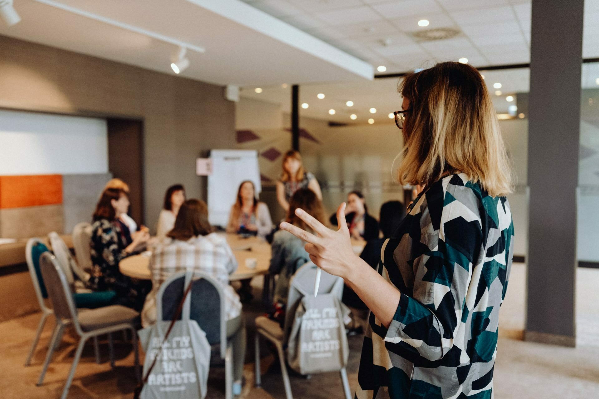 A woman speaks to a group of people sitting around a table during what looks like a meeting or workshop. She makes a peace sign with her hand. The room has modern decor with glass partitions and soft lighting, perfect for capturing moments at a photography event in Warsaw.  