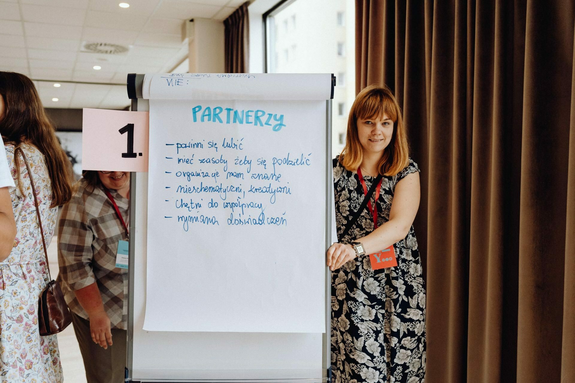 A woman in a flowery dress is standing next to a flip chart with "PARTNERS" written underneath it, with handwritten text showing the highlights in a photo essay of the event. The flip chart is marked with the number 1. In the background, other people can be seen at the window covered with curtains.  