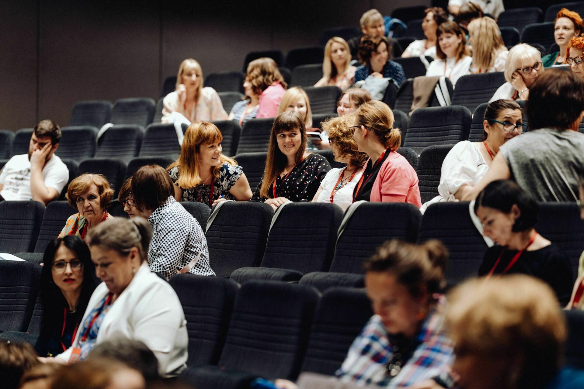 An auditorium filled with people seated and engaged in discussion. The audience, both men and women in casual attire, some talking among themselves, others listening intently or focusing on something outside the frame - ideal for event photography, where a detailed photo report of the event is captured. 