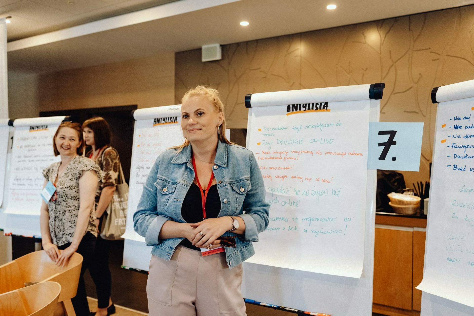 During a conference or workshop, three people stand next to large paper flip charts on which colorful captions and diagrams are displayed. One woman in the foreground is wearing a denim jacket and name badge and appears to be engaged in the activity, while the others watch and smile. This is part of our photo coverage of the event by our talented photographer for the event.  