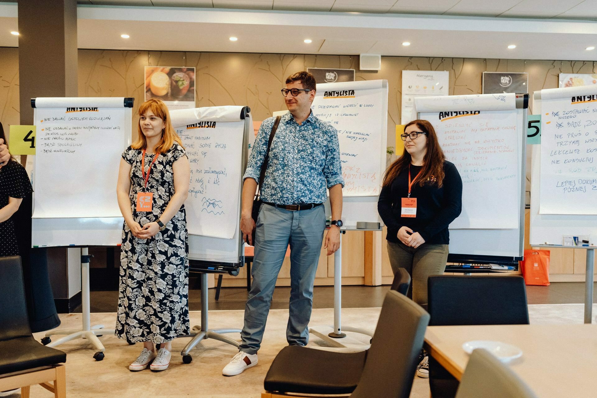 Three people stand in front of the boards in a well-lit room, creating a perfect photo-op of the event. They all have red lanyards with name badges. The woman on the left is wearing a floral dress, the man in the middle is wearing a floral shirt and glasses, and the woman on the right is wearing a black shirt and glasses.  