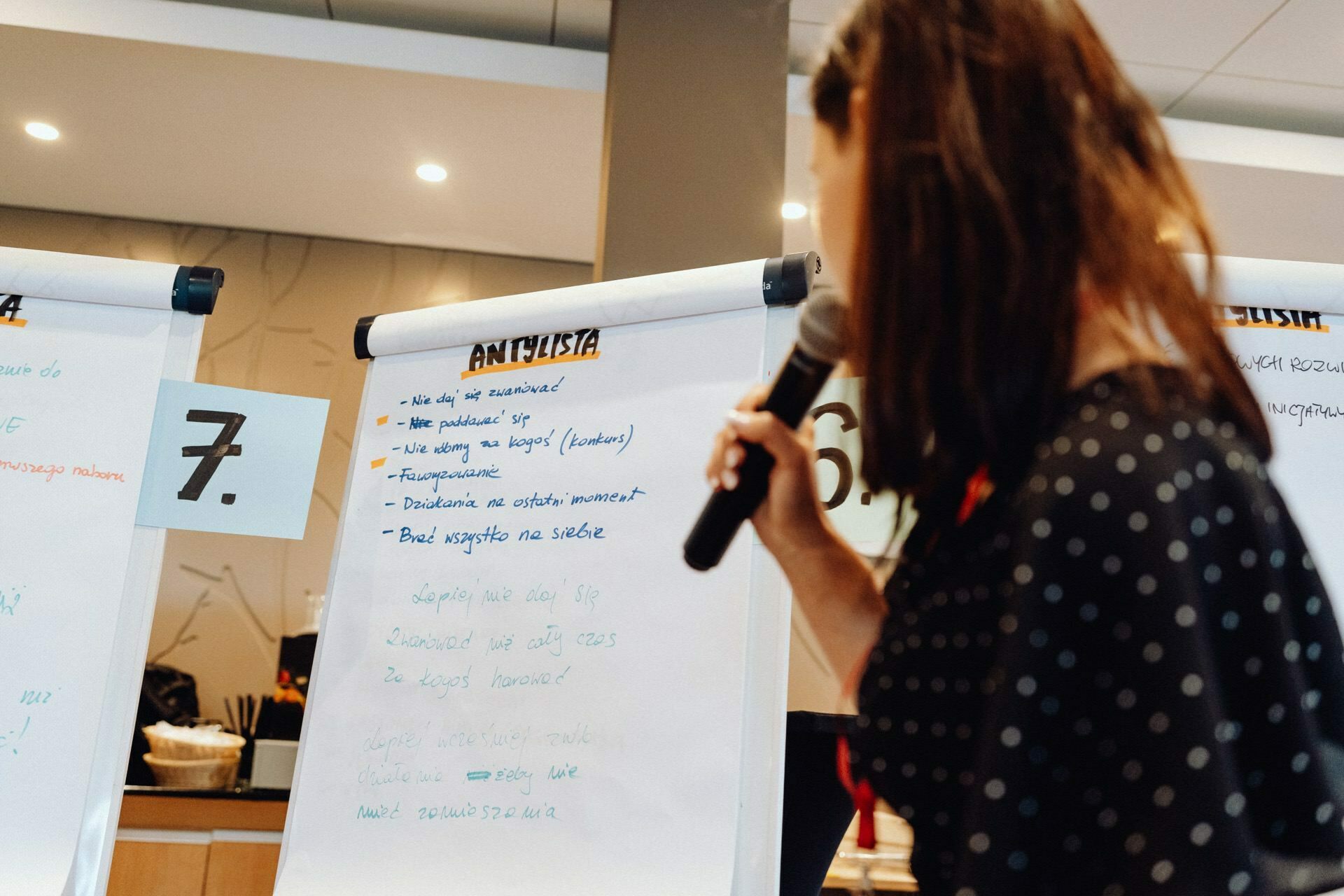 A woman holds a microphone and stands next to two flipcharts, each with handwritten notes in Polish. The flipchart on the left is numbered "7". and the one on the right, partially visible, is marked "6." The interior is characterized by office decor. This moment was captured by a photographer from Warsaw.    