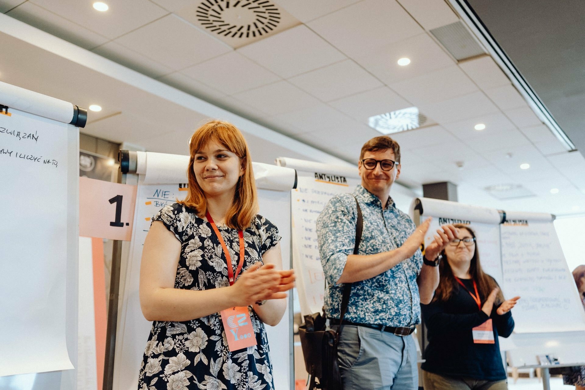 Three people are standing in a conference room, smiling and clapping. They sit in front of flip charts with various notes and papers attached. The scenery is reminiscent of a workshop or presentation, beautifully captured for a photo essay of the event. Individuals wear name badges on lanyards around their necks.   