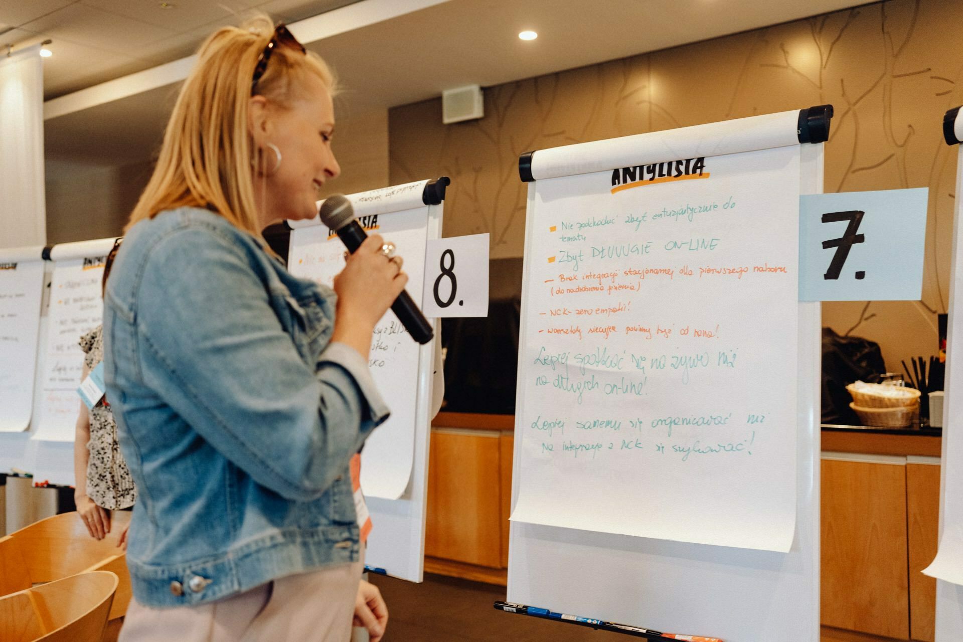 A woman holding a microphone stands in front of a flipchart with handwritten notes during an event. She appears to be presenting or discussing the content written on the chart. Two other flipcharts marked "7" and "8" are visible in the background - a moment captured by a photographer from Warsaw.  