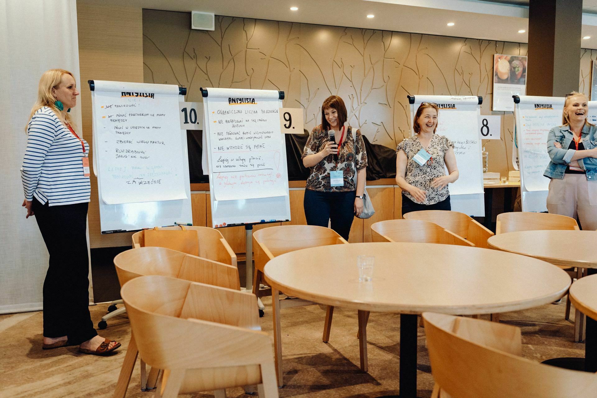 Four women stand in a conference room next to flip charts with notes. Some engage with the content on the charts. The room has round wooden tables and chairs arranged in the foreground. The atmosphere, beautifully captured by a photographer from Warsaw, gives the impression of being communal and informal.   