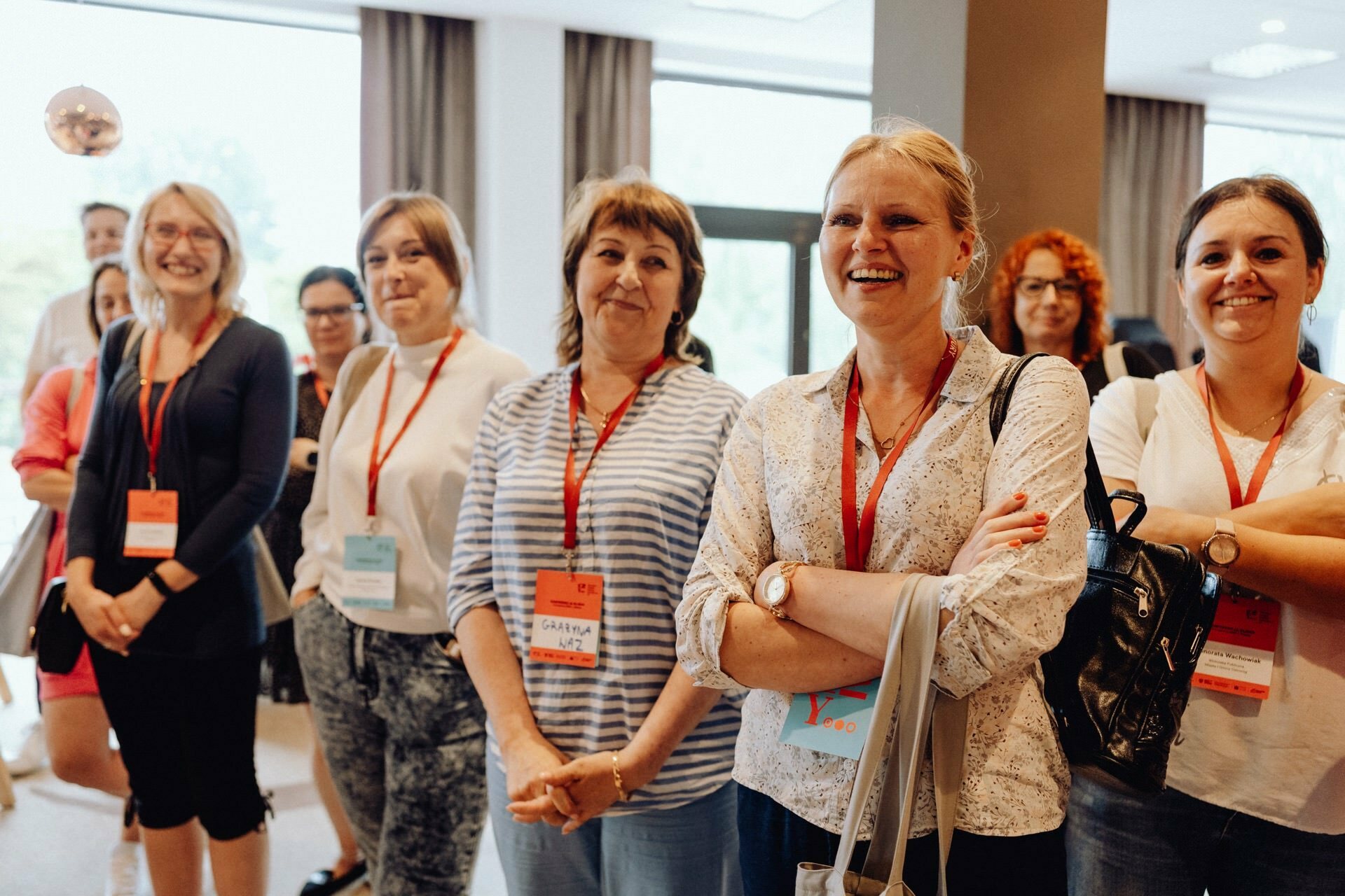 A group of seven women, all wearing name badges and lanyards, stand together in a room with large windows and curtains. Smiling and looking happy at the conference, they were beautifully captured by the photographer for the event. In the background you can see the blurry attendees mingling.  